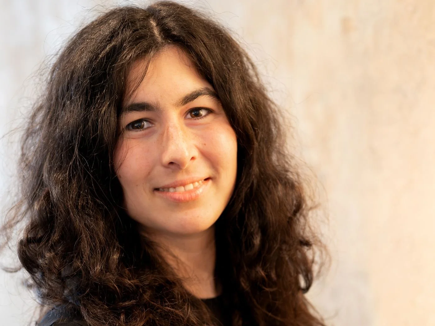 Nahaufnahme einer lachenden jungen Frau mit langen, lockigen braunen Haaren vor einem neutralen Hintergrund.