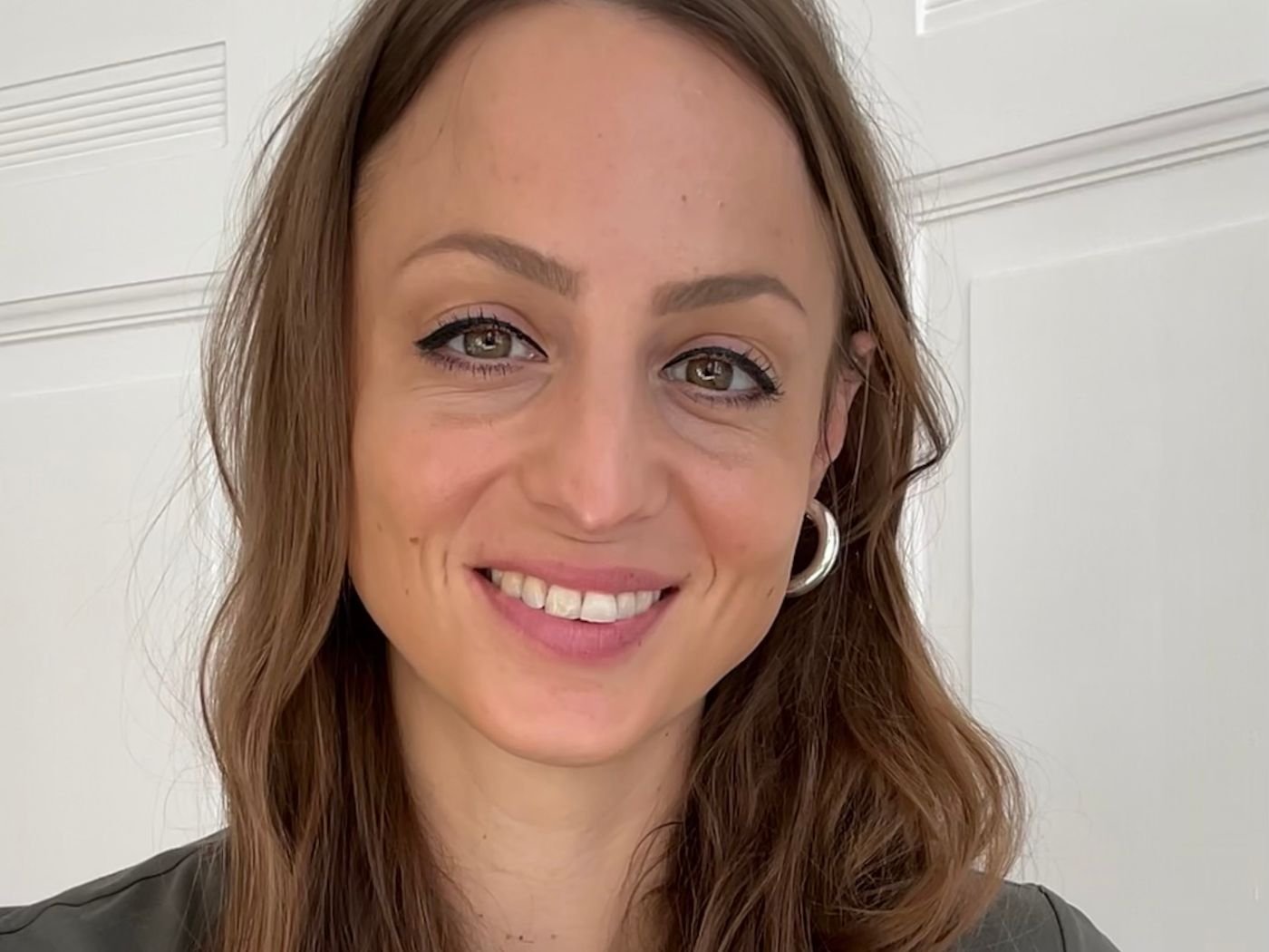 Lächelnde Frau mit braunen lockigen Haaren, hellen Augen, silberner Ohrstecker, vor weißer Tür.