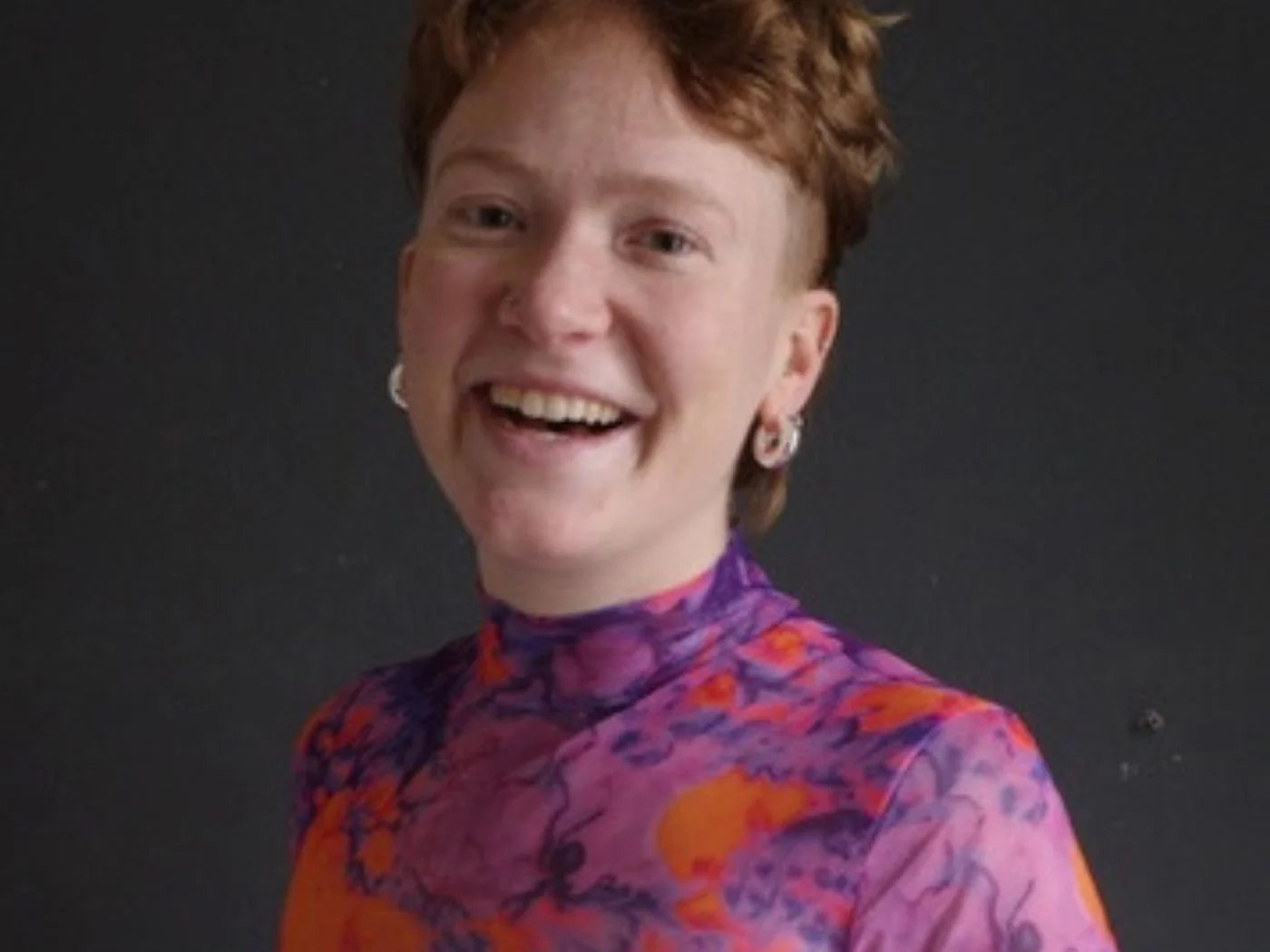 Lächelnde Person mit Kurzhaarfrisur und buntem, grafischem Oberteil vor dunklem Hintergrund.