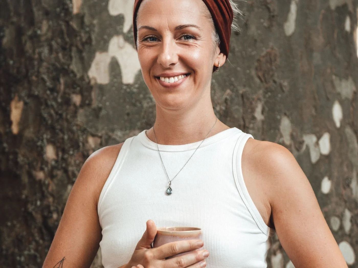 Lächelnde Frau mit roten Haaren, trägt weiße ärmellose Bluse, stilvolle Halskette, hält Tasse, steht vor einer Steinmauer.