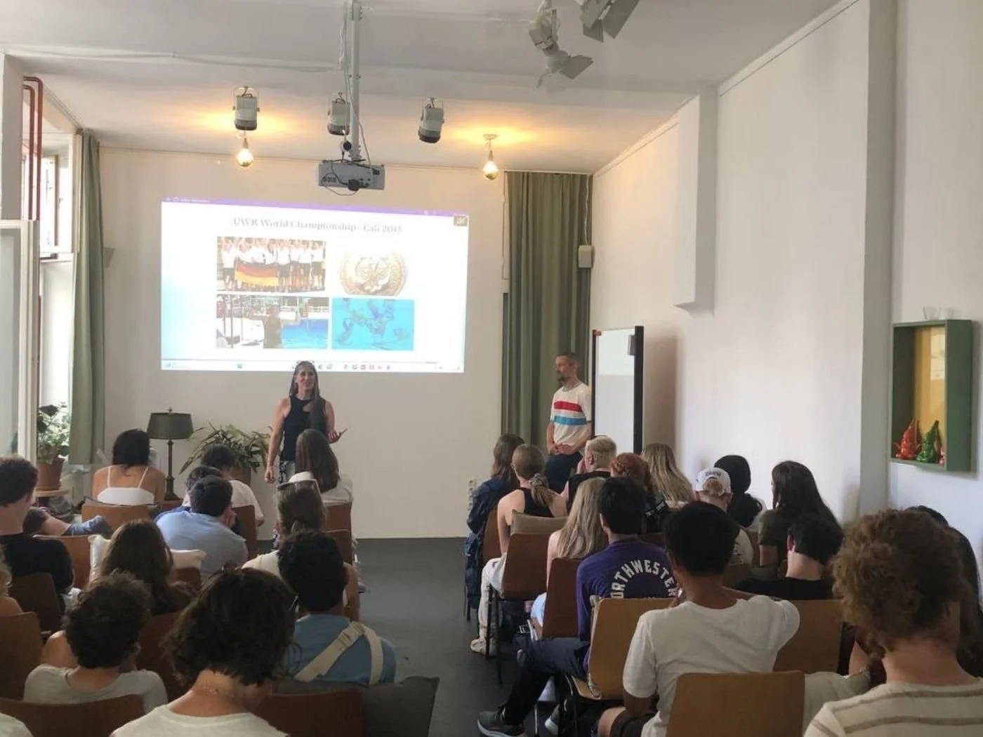 Ein Vortragssaal mit mehreren Menschen, die einem Vortrag zuhören. An der Wand eine Präsentationsfolie mit Bildern und Text, zwei Menschen stehen vorne bei der Präsentation.