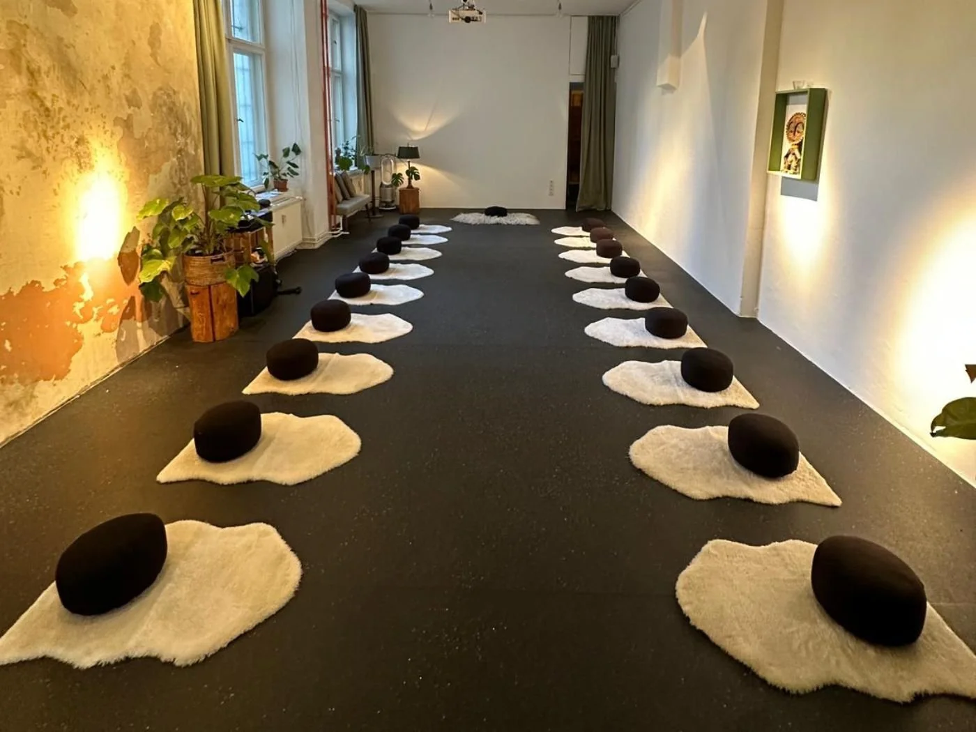 Ein Raum mit Unterlagen und schwarzen Kissen auf weißen Fellmatten, wahrscheinlich für eine Meditations- oder Yogastunde, mit Pflanzen und Dekorationen.