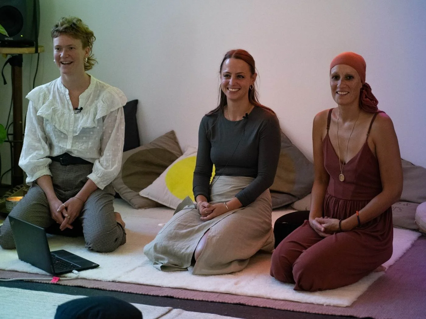 Drei Frauen sitzen auf einem Teppich vor einem Kissen und einem Laptop, sie lächeln und scheinen an einem Gespräch oder einer Präsentation teilzunehmen.