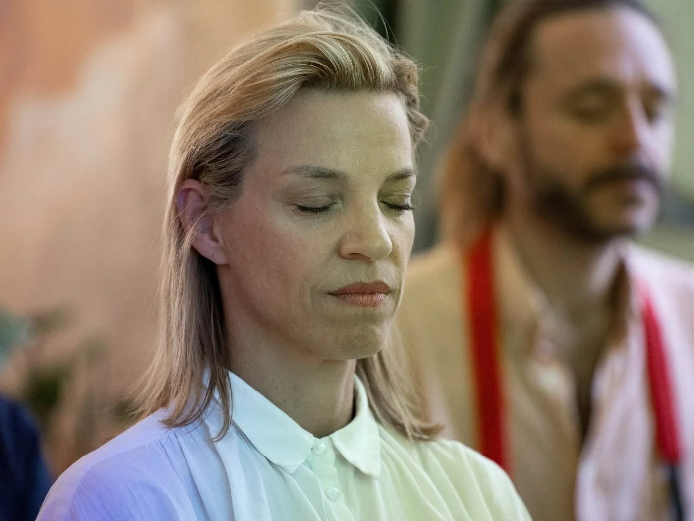 Zwei Menschen, eine blonde Frau in weißem Hemd und eine dunkle Frau mit glattem Haar und rotem Träger-Top, stehen mit geschlossenen Augen in einer entspannten Haltung.