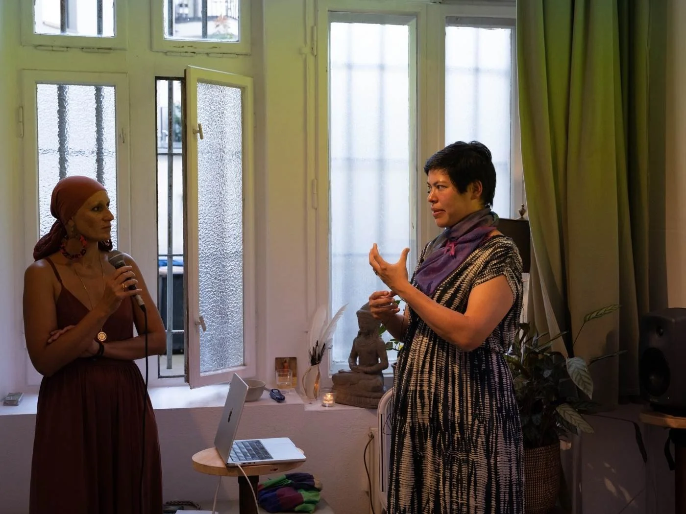 Zwei Frauen sprechen in einem Raum, eine mit Mikrofon, die andere gestikulierend, vor einem offenen Fenster mit Vorhängen.