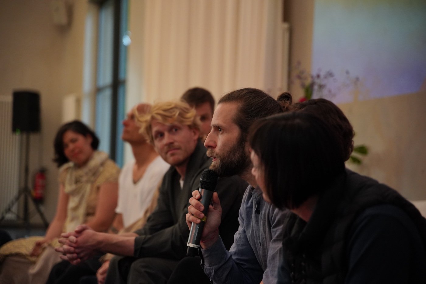 Menschen sitzen in einer Reihe und sprechen bei einer Veranstaltung, eine Person hält ein Mikrofon.