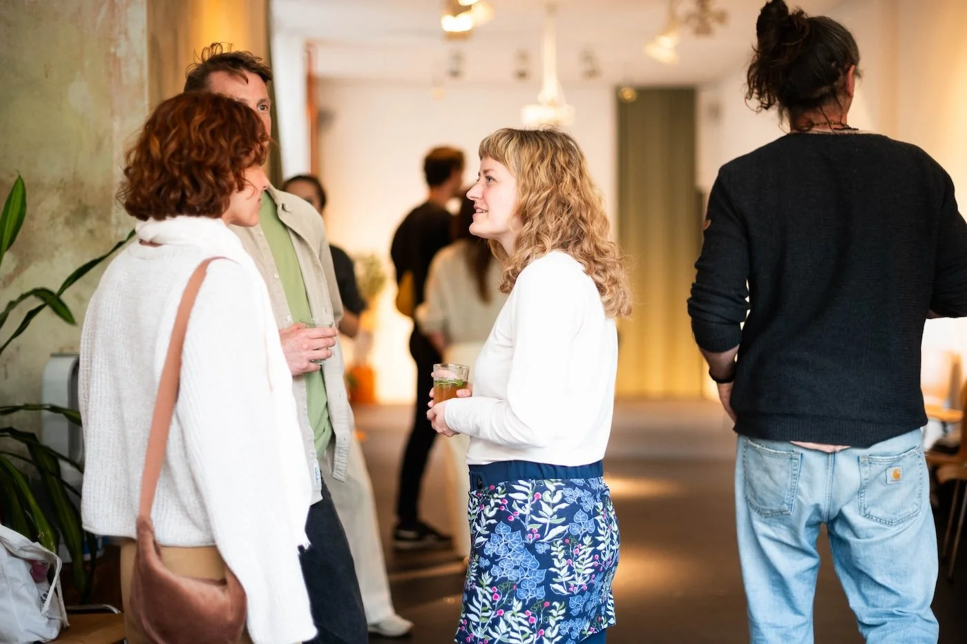 Menschen beim Gespräch in einem Raum, manche halten Getränke, im Hintergrund weitere Personen, geschäftiges Treffen.