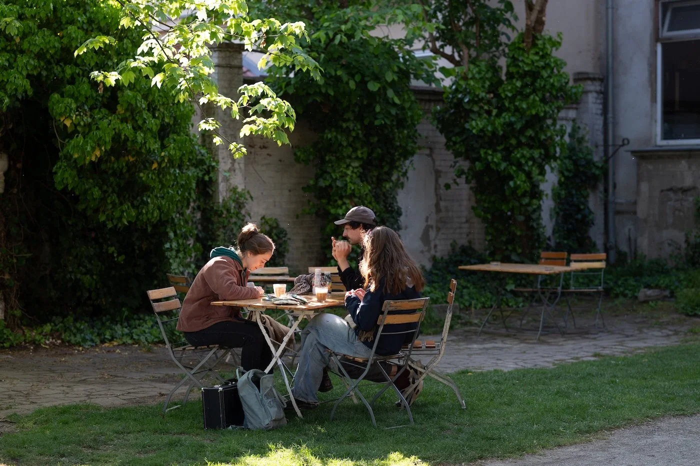 Drei Menschen sitzen an einem kleinen Holztisch im Freien, bei Sonnenlicht, mit Getränken und Karten, vor einem Gebäude mit Pflanzen und Bäumen im Hintergrund.