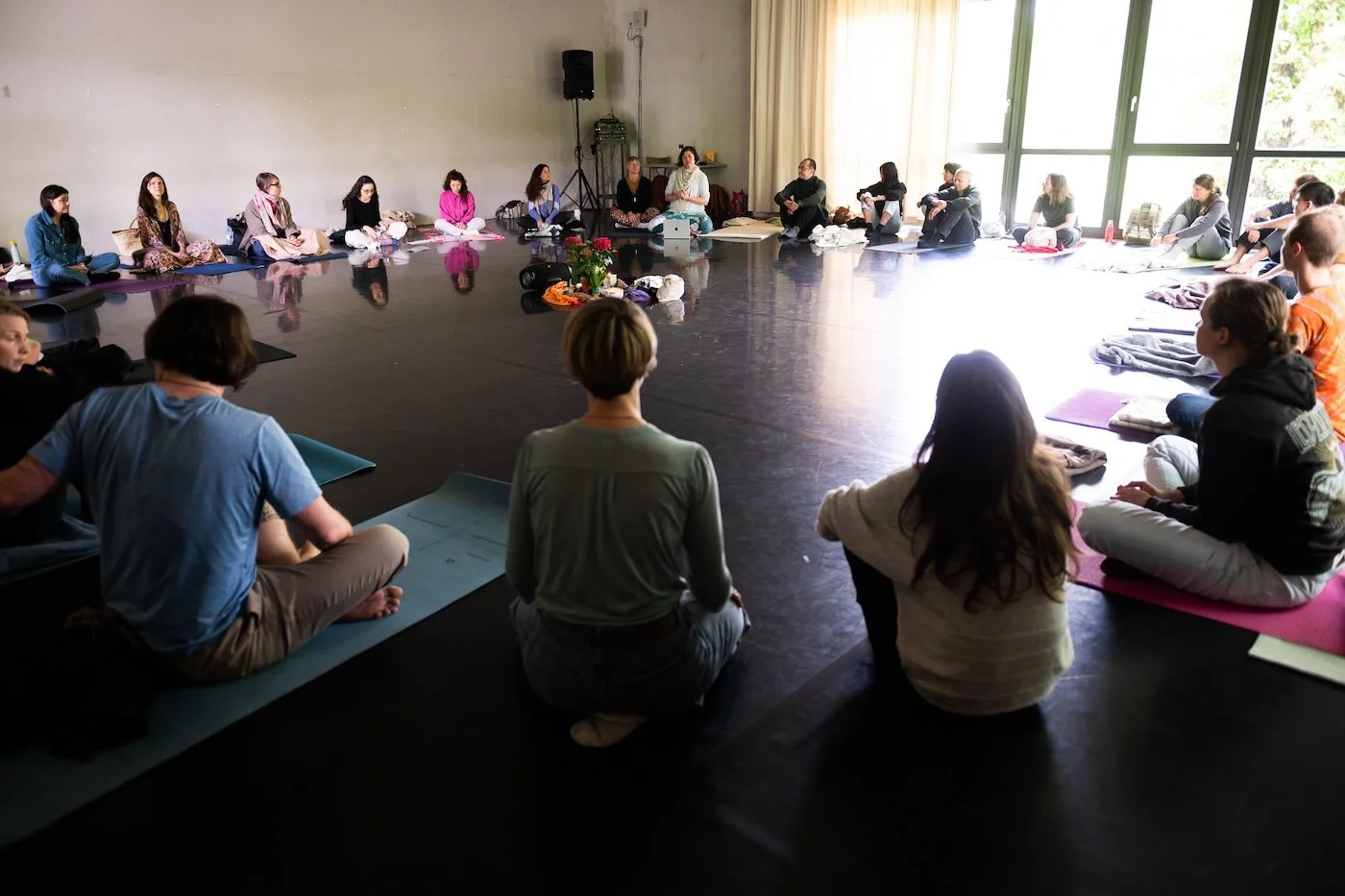 Menschen sitzen im Kreis auf Yogamatten in einem großen, hell erleuchteten Raum, vermutlich bei einem Meditations- oder Yoga-Workshop.