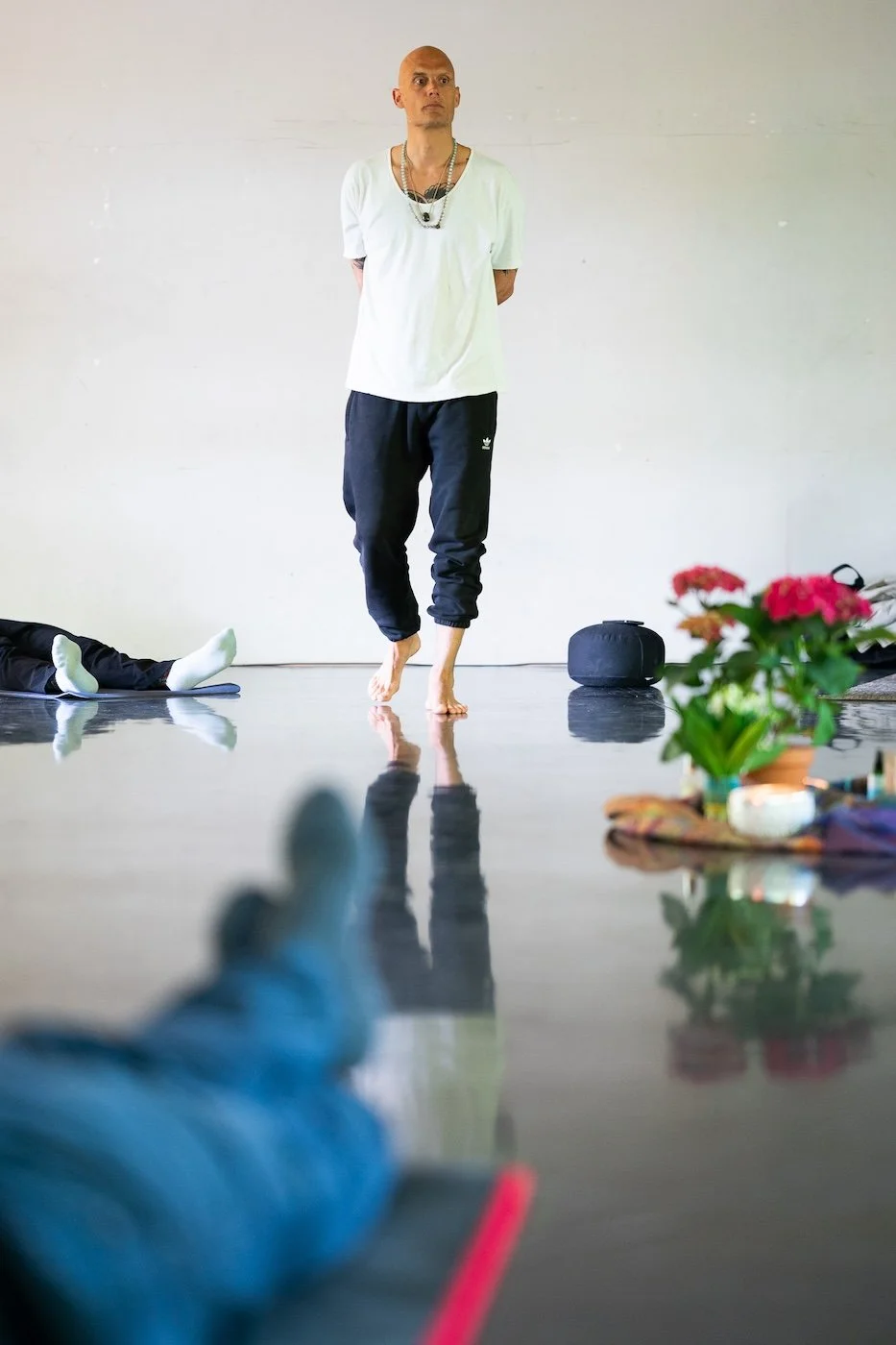 Ein Mann tanzt barfuß in einem Raum, während eine Person im Hintergrund auf dem Rücken liegt. Im Vordergrund ist eine Hand mit dicken, blauen Kleidung sichtbar, und im unteren rechten Bereich gibt es einen Blumentopf mit roten Blumen.