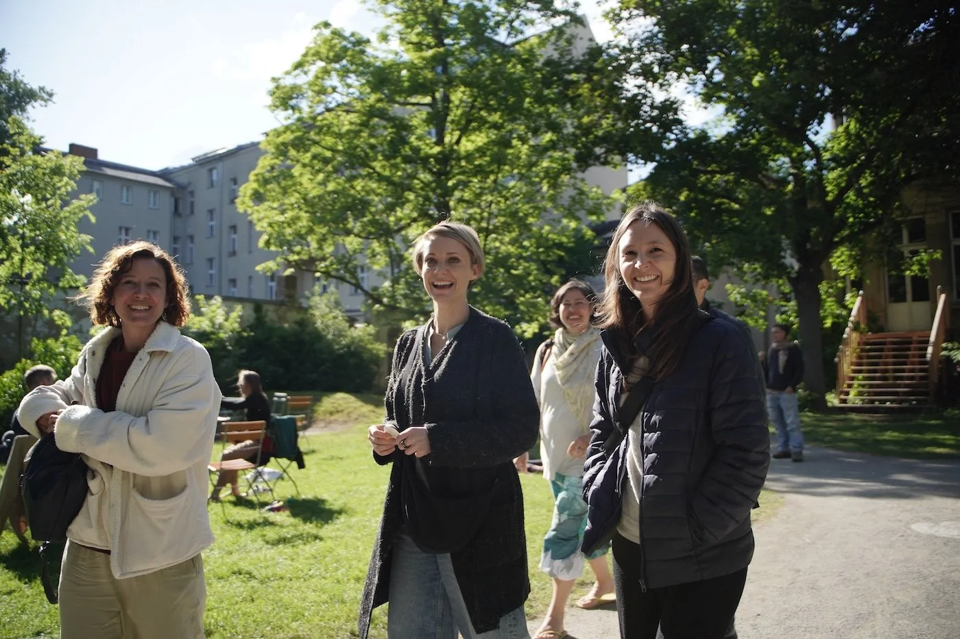 Vier lachende Frauen stehen im Park bei sonnigem Wetter, im Hintergrund Bäume und Gebäude, andere Personen sitzen auf Bänken.