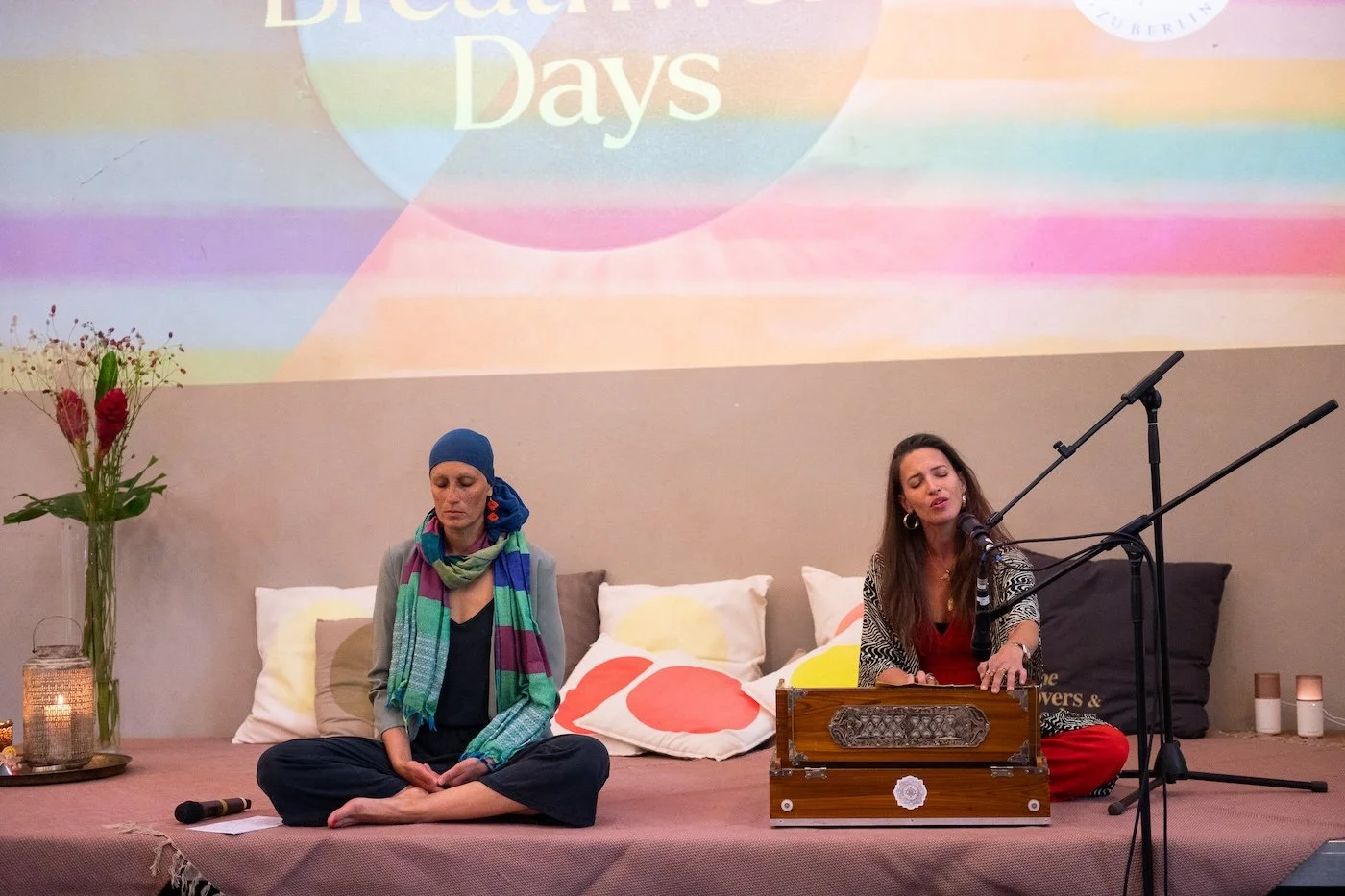 Zwei Frauen sitzen auf einem Bühnenpodest, eine spielt eine Harmonium, die andere sitzt im Meditationssitz mit geschlossenen Augen. Im Hintergrund eine große Projektionswand mit buntem 'Dream Days'-Logo. Dekoratives Element auf der linken Seite ist e
