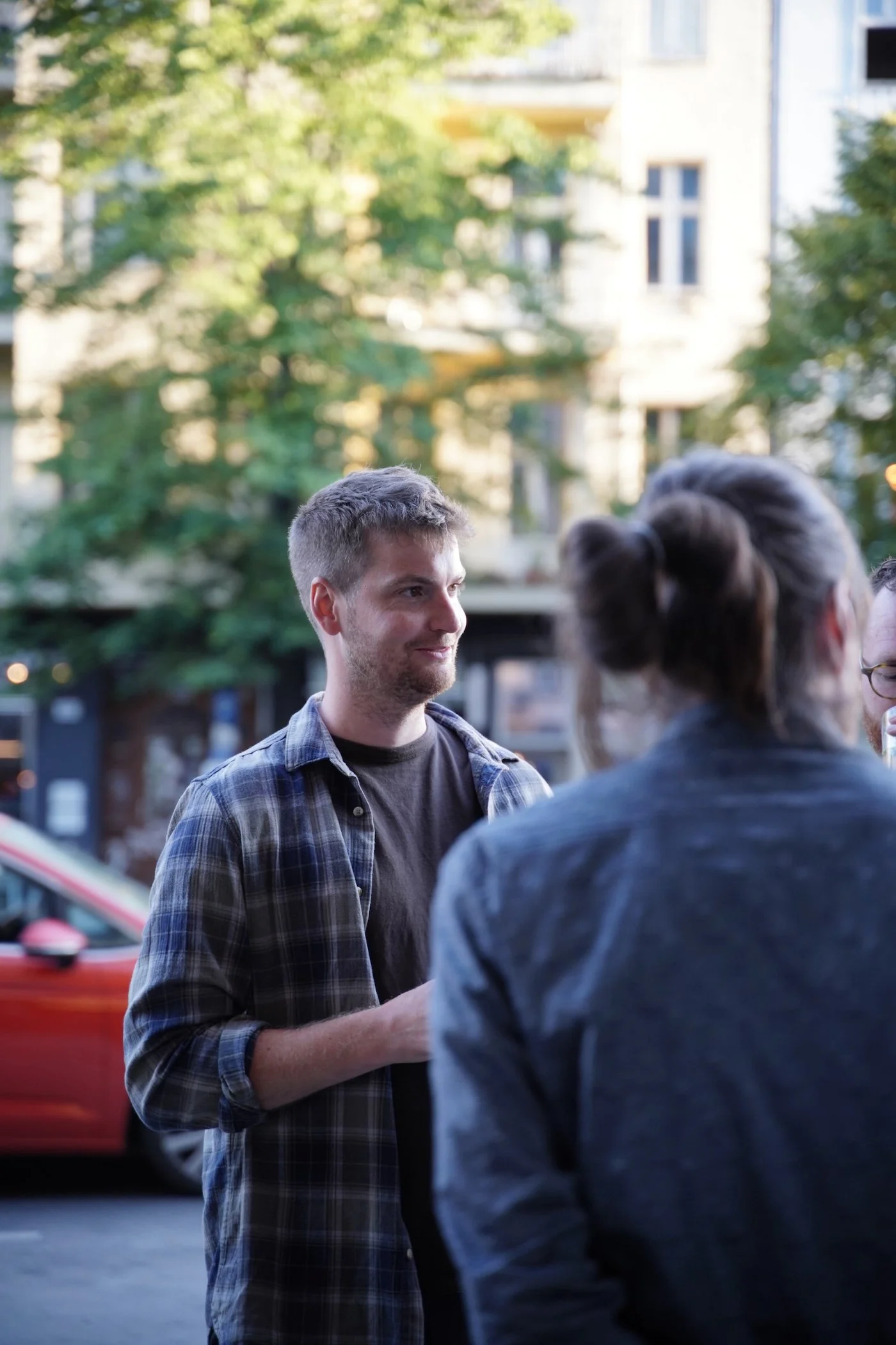 Drei Menschen sprechen im Freien in einer Stadt bei Sonnenuntergang, im Hintergrund Bäume und Gebäude.