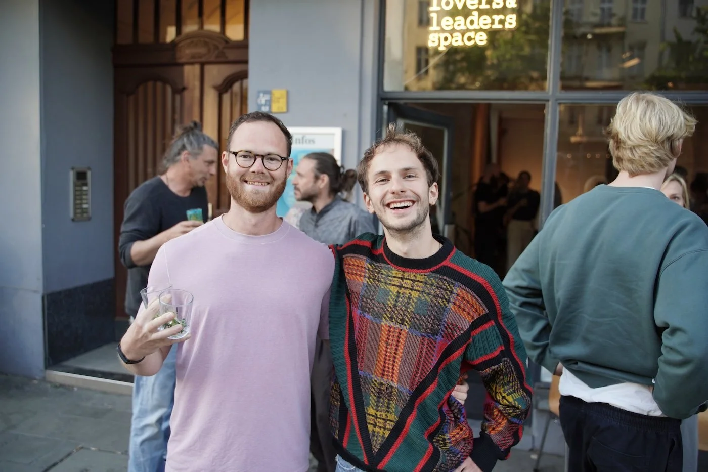 Zwei lachende Männer stehen draußen vor einem Café oder Restaurant, umgeben von weiteren Personen unter Abendlicht.