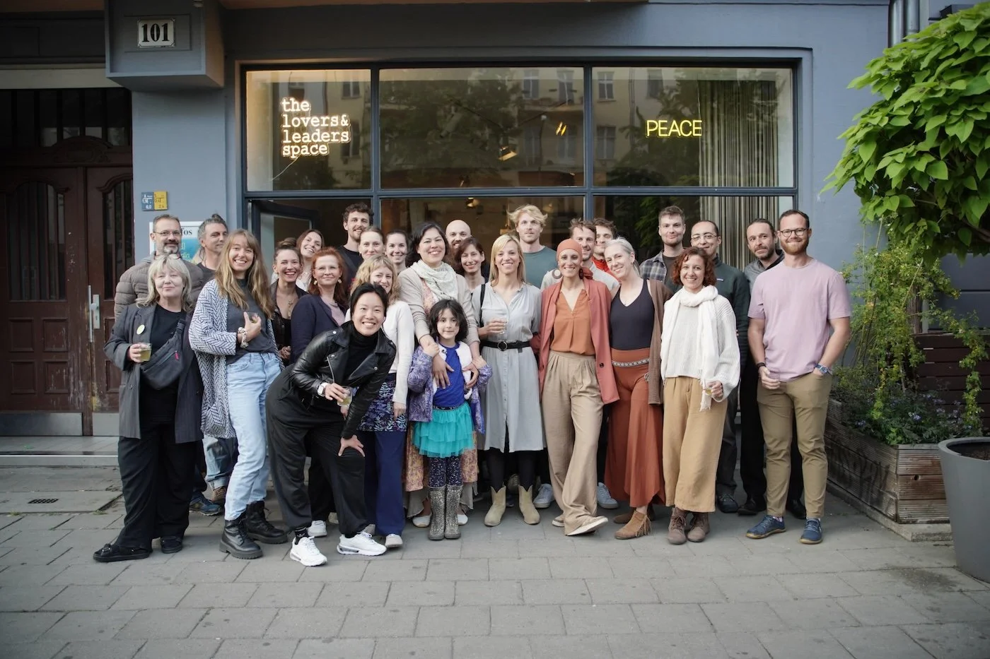 Eine Gruppe von etwa 20 Menschen steht vor einem Gebäude mit der Nummer 101. Sie posieren für ein Gruppenfoto und wirken fröhlich. Im Hintergrund sind Schaufenster mit Neon-Schildern, die liest: "the lovers & leaders space" und "PEACE".