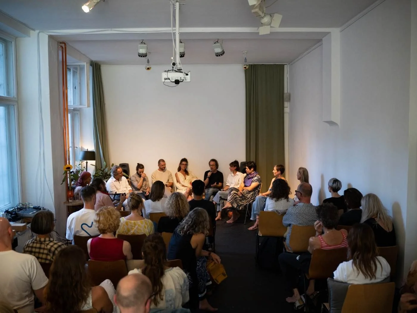 Eine Gruppe von Menschen sitzt in einem kalten weißen Raum und hört einer Diskussion zu, die auf einem kleinen Podium stattfindet.