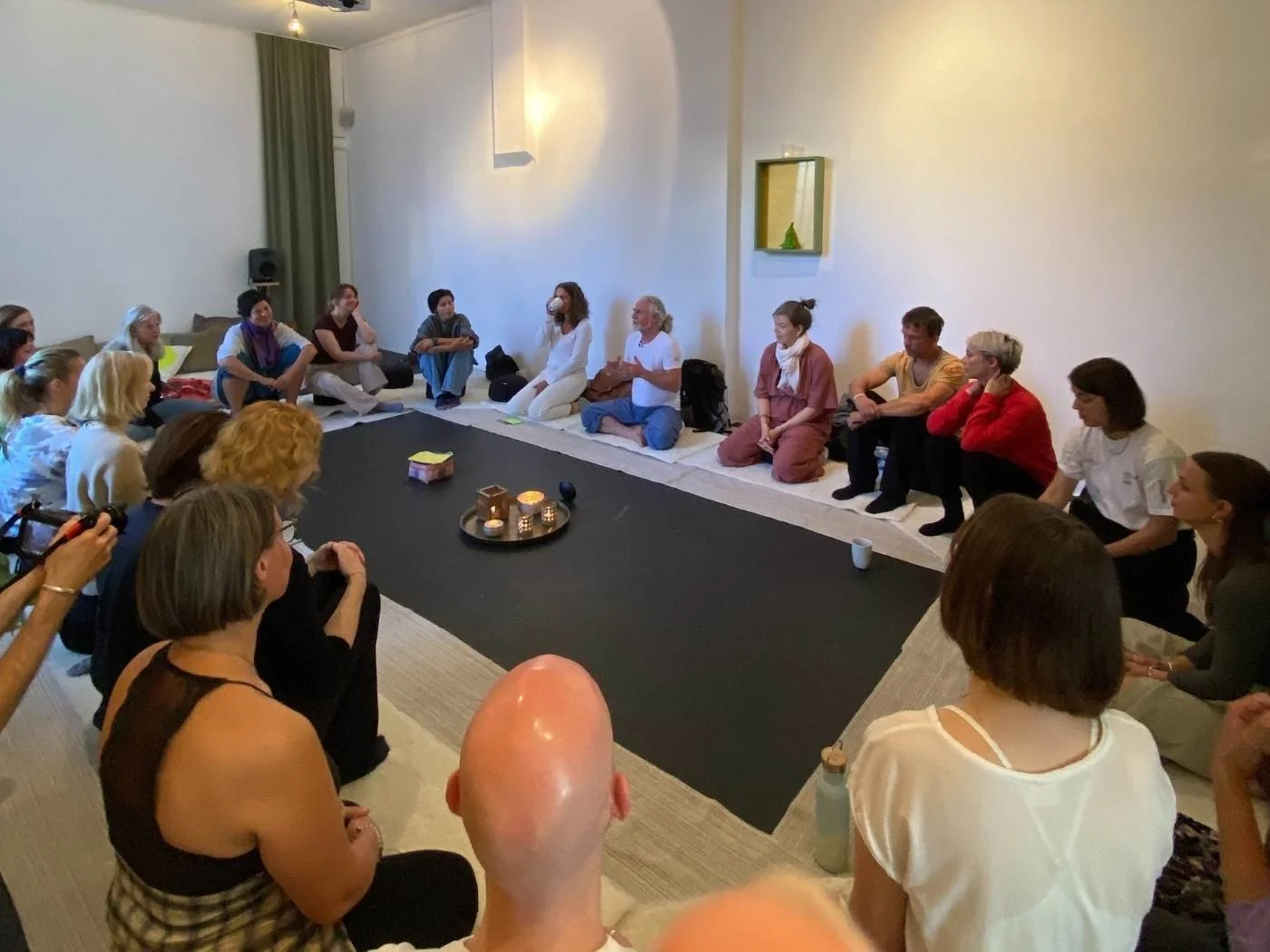 Menschen sitzen in einem Kreis auf dem Boden eines Zimmers, das für eine Gruppeaktivität oder Meditation arrangiert ist, mit Kerzen und einer kleinen Dekoration in der Mitte.