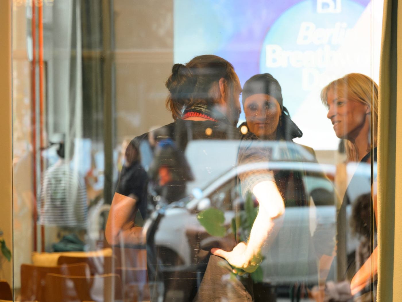 Drei Frauen sprechen sich in einem Café durch eine Fensterfront ab, mit reflektiertem Auto und Passanten im Hintergrund.