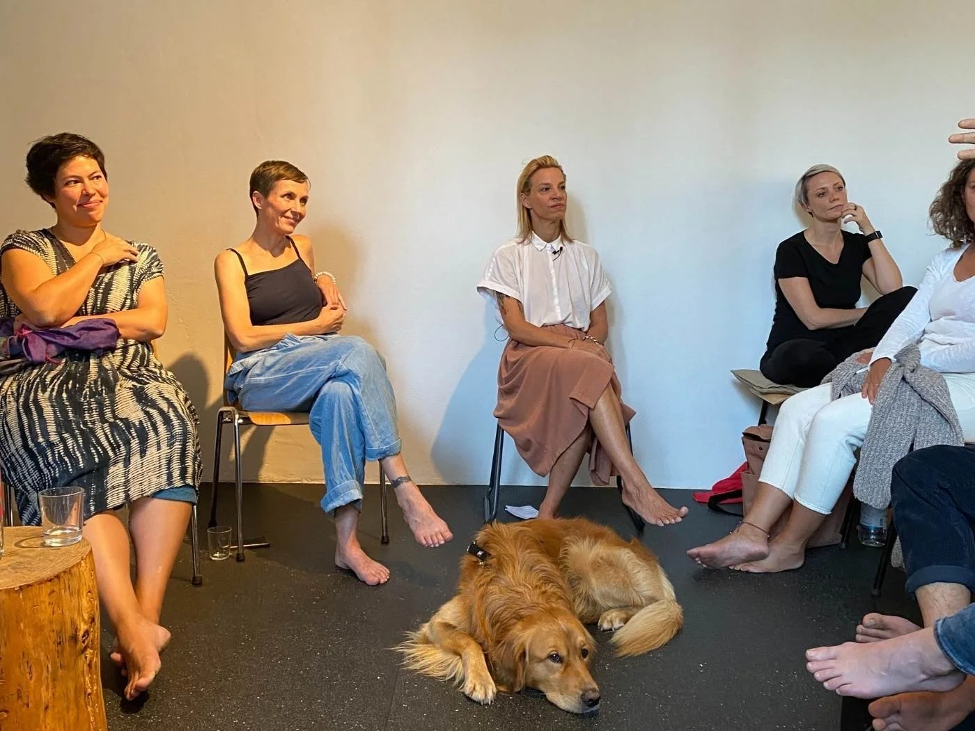 Fünf Frauen sitzen in einem Raum, die eine Frau spricht und der Hund liegt auf dem Boden. Die Frauen sind in verschiedenen Farben gekleidet, einige sind barfuß.