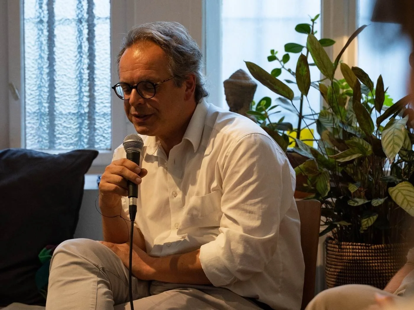 Ein älterer Mann mit Brille trägt ein weißes Hemd und hält ein Mikrofon in der Hand, während er spricht. Im Hintergrund sind Pflanzen und eine Fensterfront sichtbar.