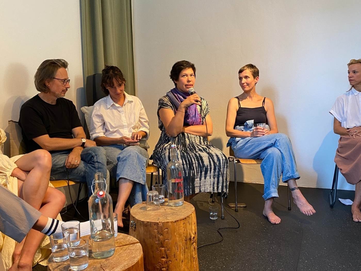 Mehrere Menschen sitzen auf Stühlen in einem Raum und nehmen an einer Diskussion oder einem Panel teil, eine Frau spricht mit einem Mikrofon.