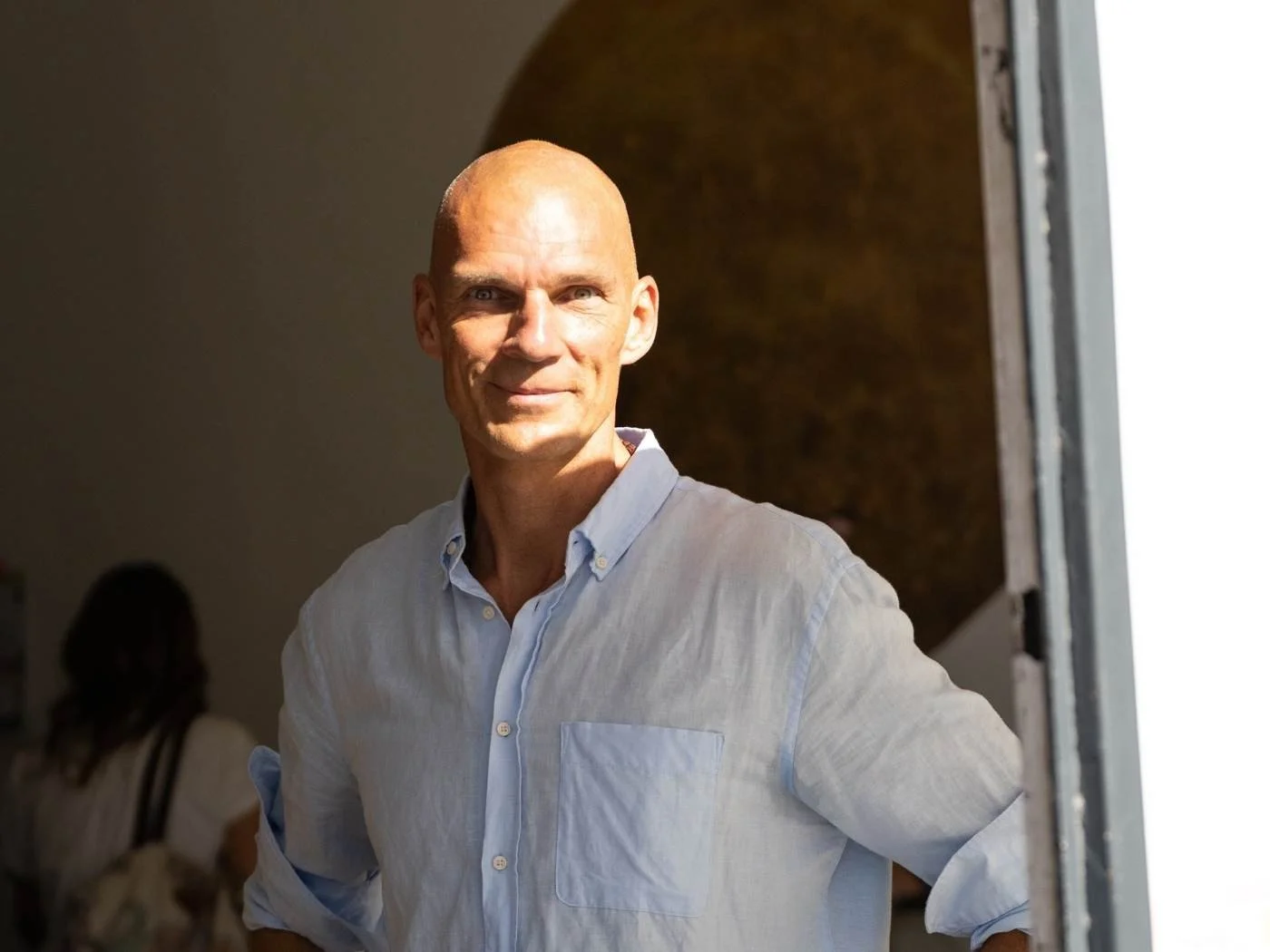 Mann mit blauer Hemd, lächelnd, steht bei einer Tür in einem Raum mit dunklem Hintergrund und Licht, das auf sein Gesicht fällt.