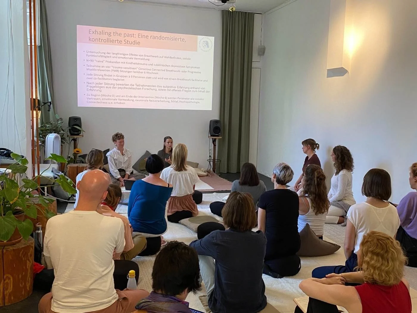 Menschen sitzen im Kreis in einem Raum und schauen auf eine Projektionswand, auf der eine Präsentation zu sehen ist.