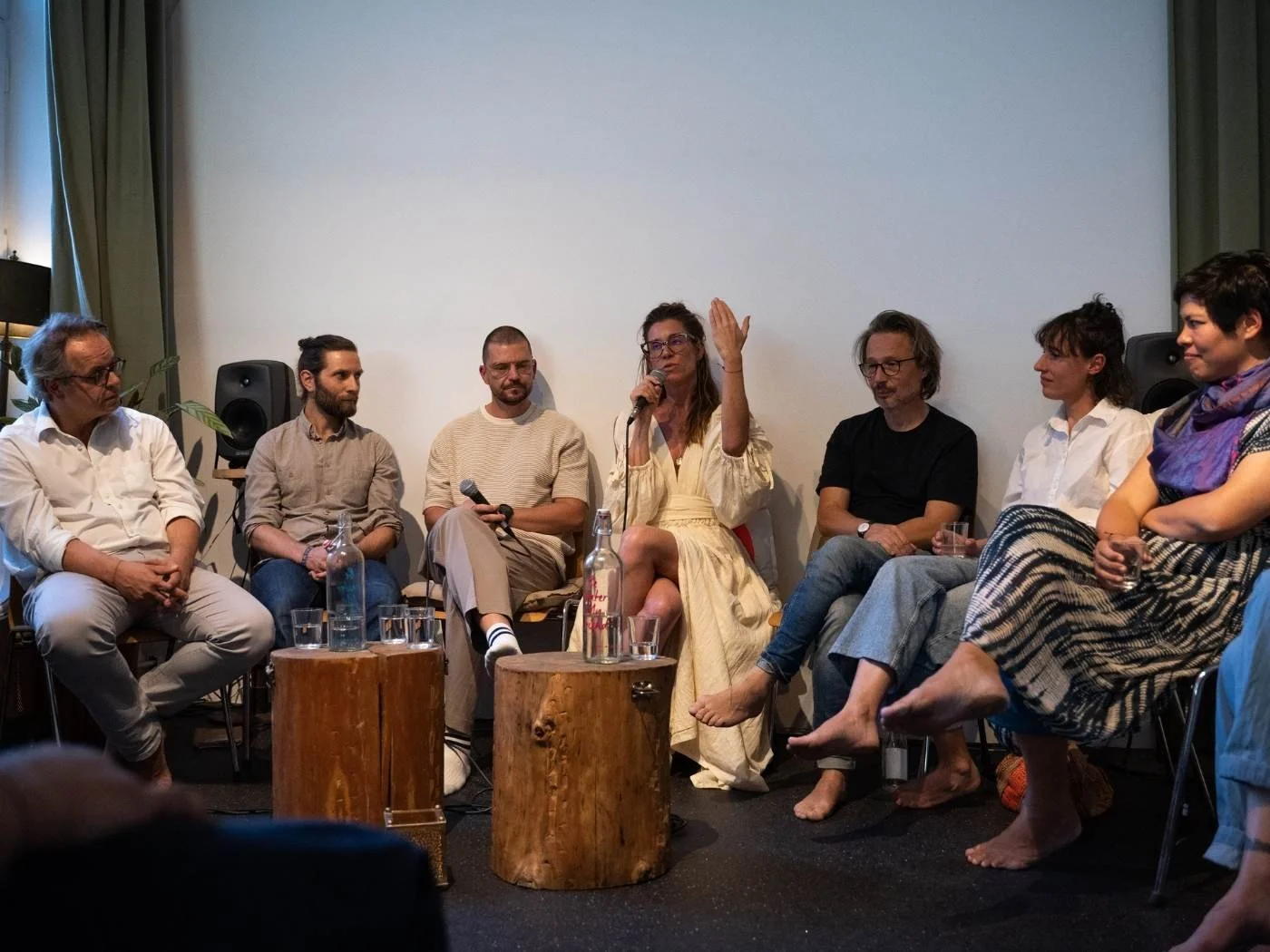 Eine Gruppe von Menschen sitzt in einer Runde bei einer Diskussion, einige halten Mikrofone, im Hintergrund ein weißer Wand, vor ihnen steht ein Holzblock mit Wasserflasche und Gläsern.