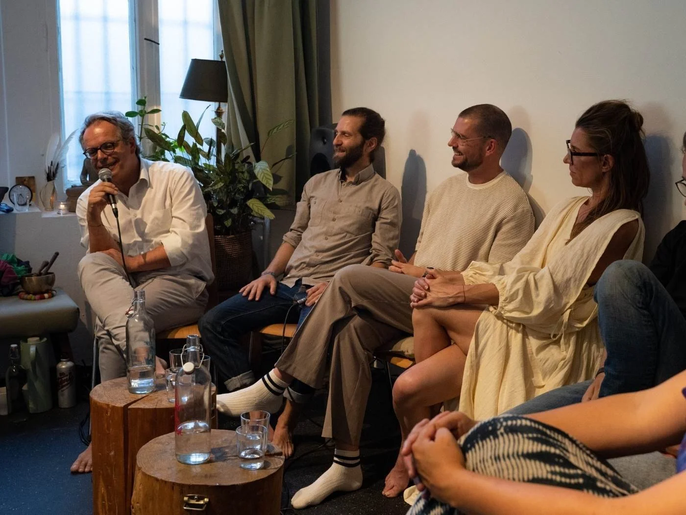 Menschen sitzen in einer gemütlichen Runde und lachen bei einer Diskussion oder einem Gespräch, in einem Raum mit Pflanzen und Fenstern im Hintergrund.