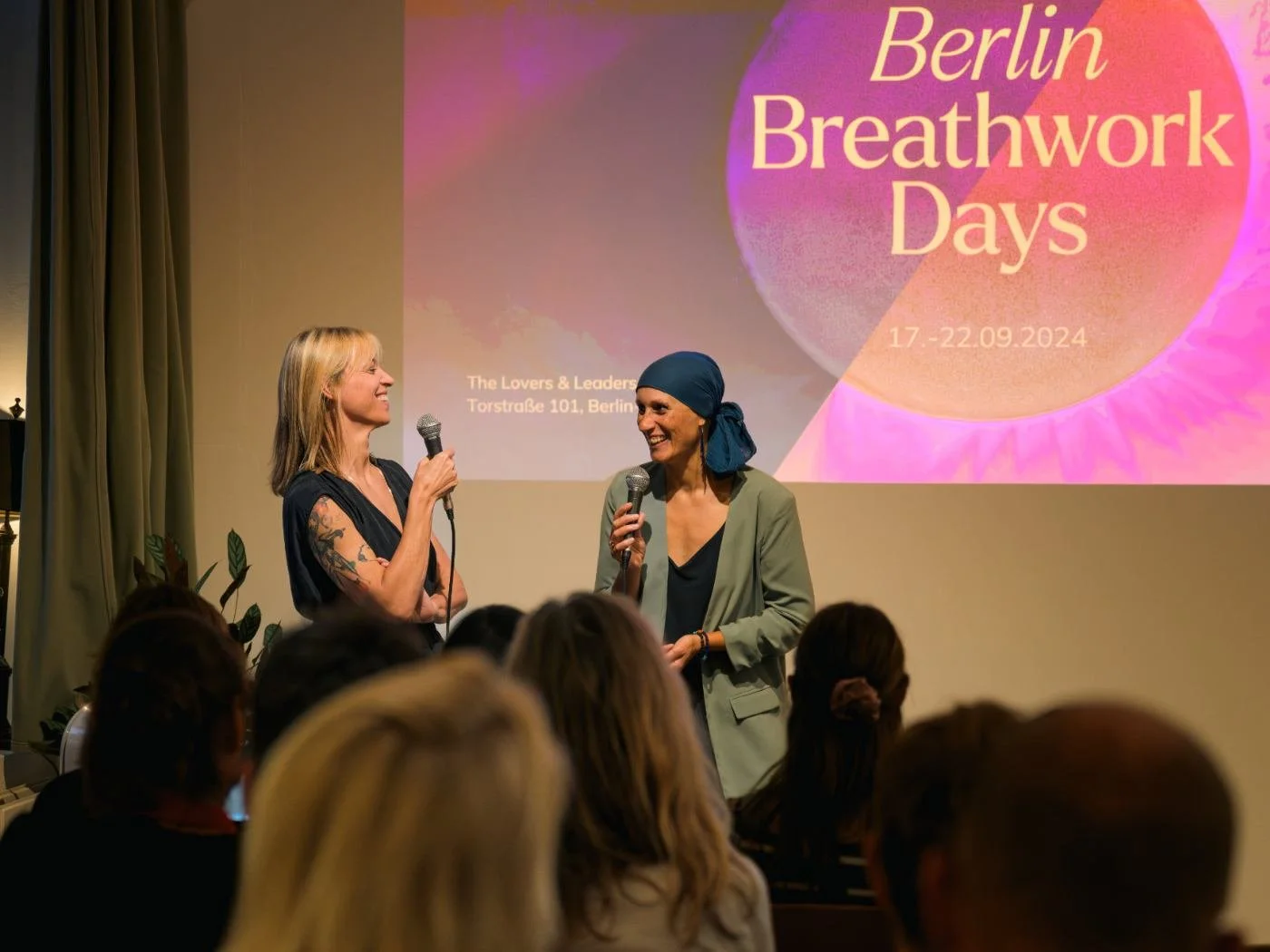 Zwei Frauen sprechen auf einer Bühne während eines Events namens 'Berlin Breathwork Days', das vom 17. bis 22. September 2024 stattfindet, vor einem großen Bildschirm, auf dem die Veranstaltungshinweise zu sehen sind. Das Publikum sitzt im Vordergrun