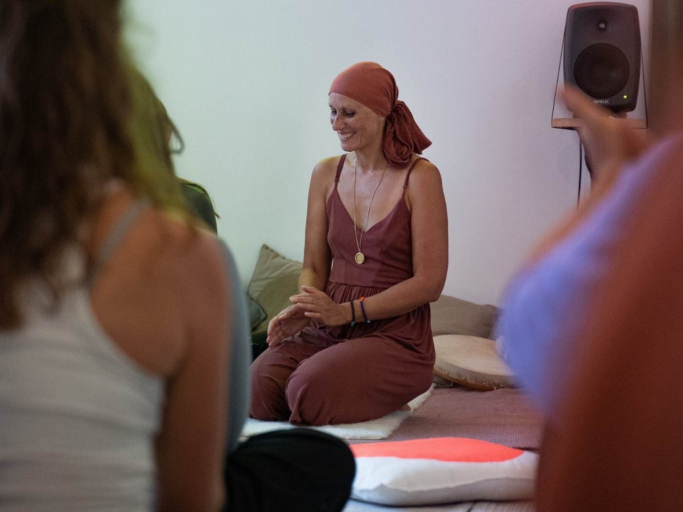 Eine Frau in einem rotbraunen Kleid und einem roten Kopftuch ist beim Meditieren oder Gebet, sitzend auf einem Kissen in einer ruhigen Ecke eines Raumes, umgeben von mehreren Menschen.