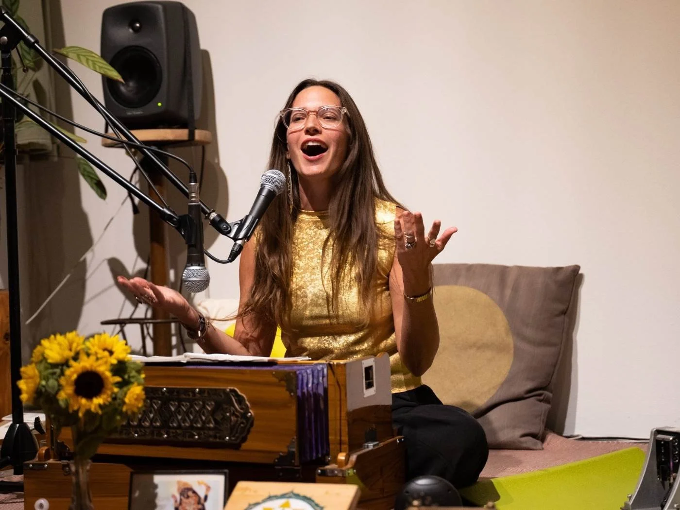 Eine Frau singt oder spricht vor einem Mikrofon in einem Raum, umgeben von Musik- und Dekorationsgegenständen.