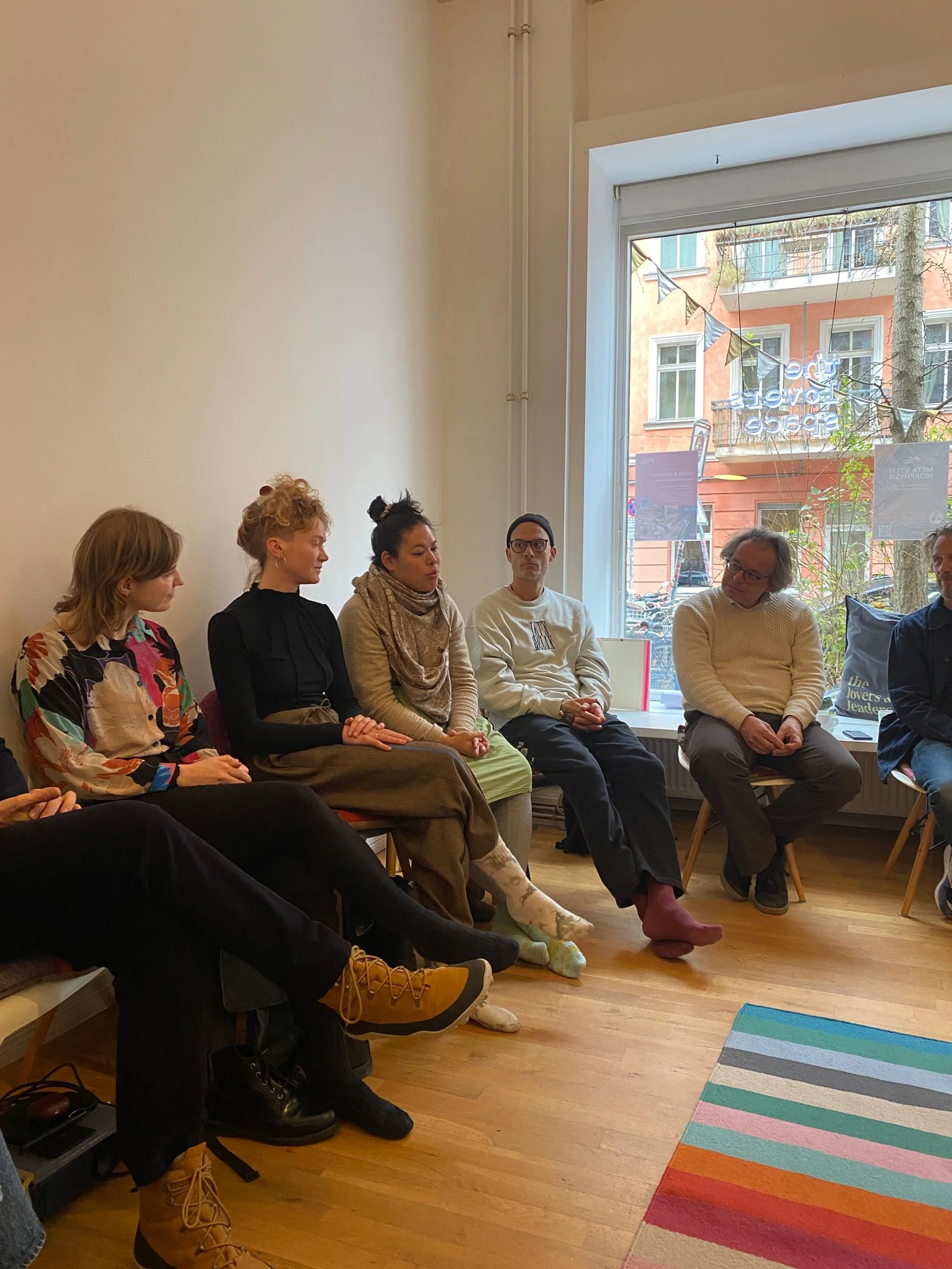 Sechs Menschen sitzen in einem Raum an einem runden Treffen, die an einem Gespräch teilnehmen. Sie sitzen auf Stühlen neben einem großen Fenster, das auf eine Straße mit bunten Gebäuden blickt.