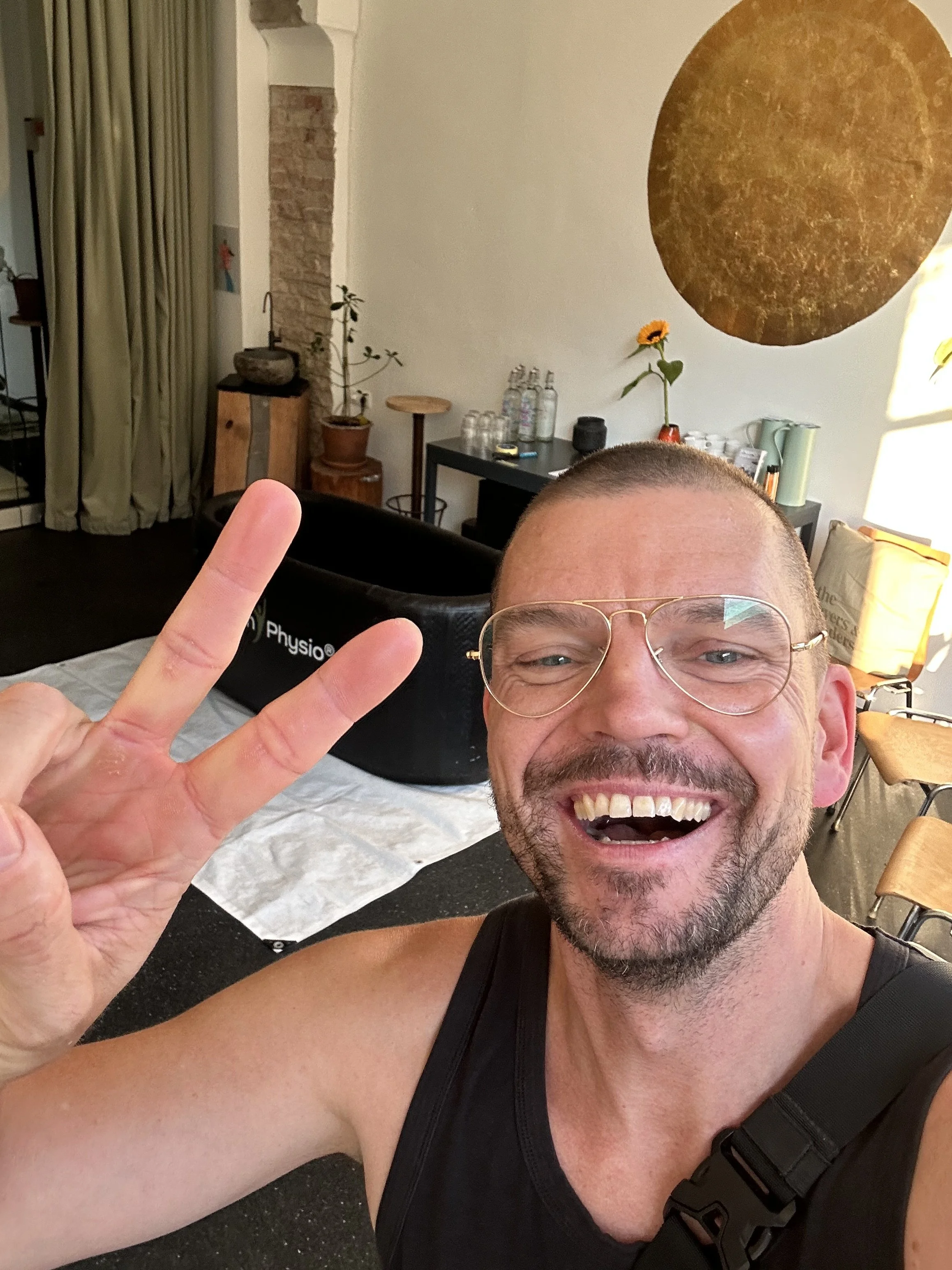 Lächelnder Mann mit Brille macht Peace-Zeichen in einem Raum mit Trainingsgerät und Pflanzen im Hintergrund.