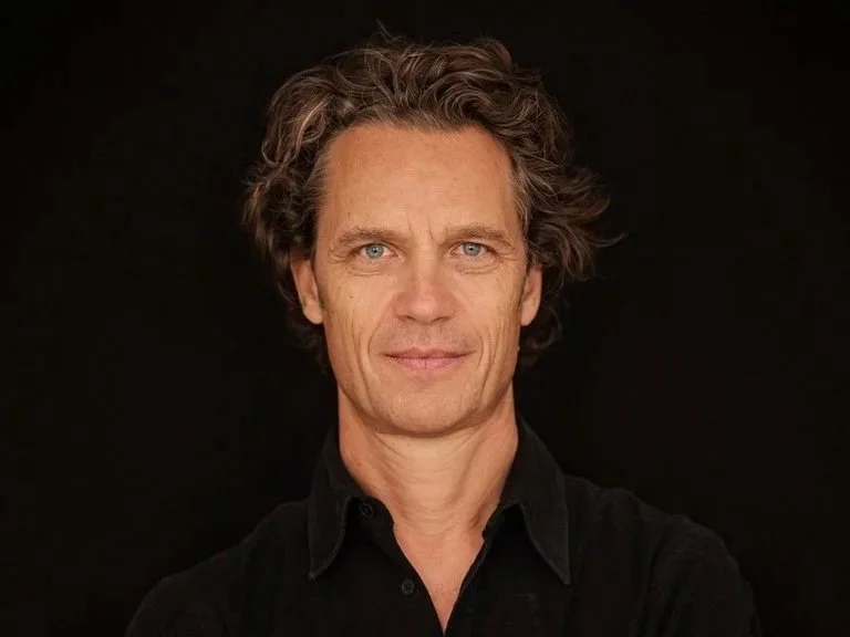 Porträt eines Mannes mit dunkelblondem, leicht gewelltem Haar, blauen Augen und hellem Teint vor schwarzem Hintergrund.