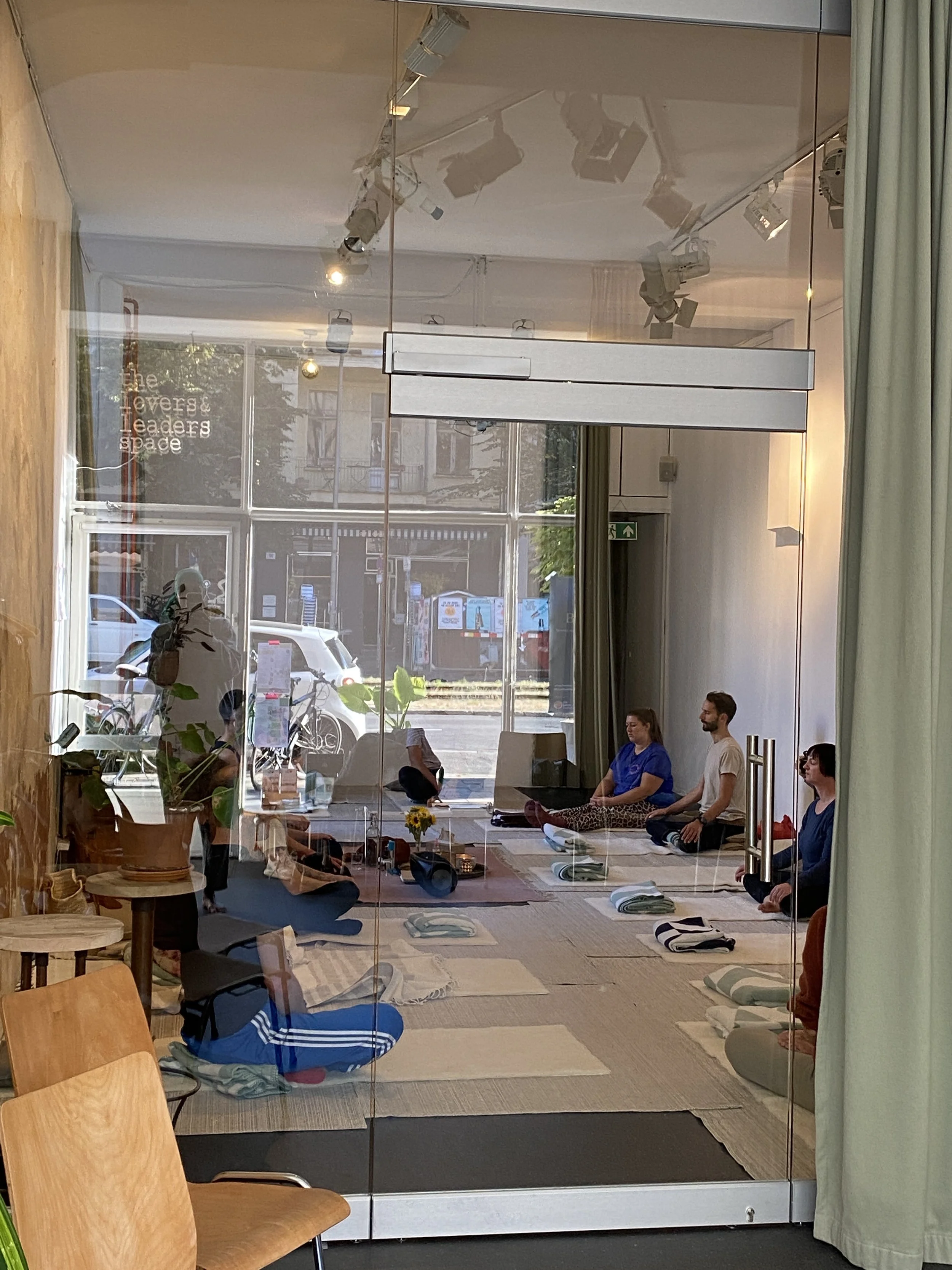 Menschen, die in einem Raum auf Yoga-Matten eine Meditation machen, durch eine Glasscheibe betrachtet.