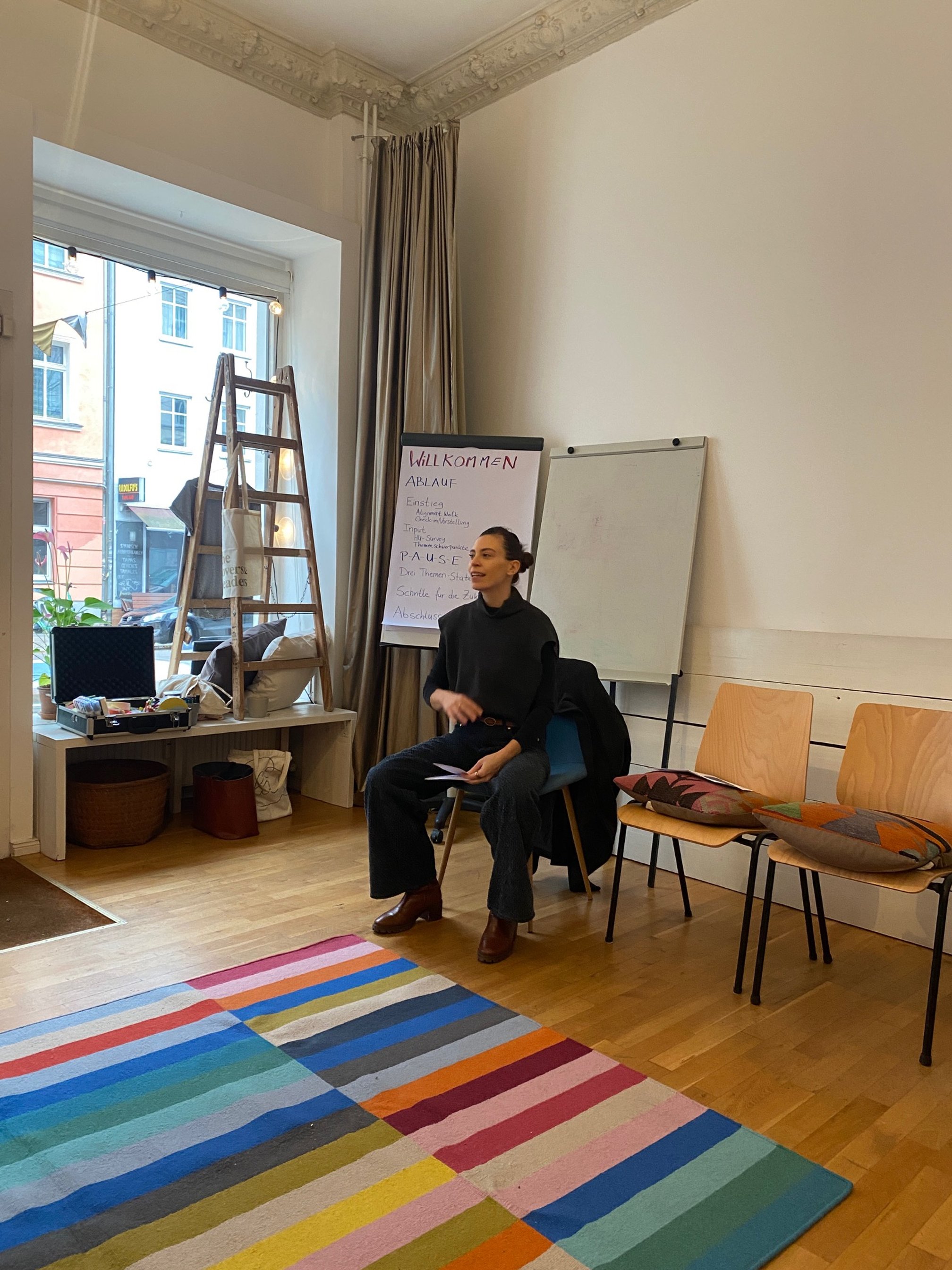 Eine Frau sitzt in einem Raum vor einer Whiteboard und einem Flipchart, es scheint eine Präsentation oder ein Treffen zu sein, mit buntem Teppich im Vordergrund.