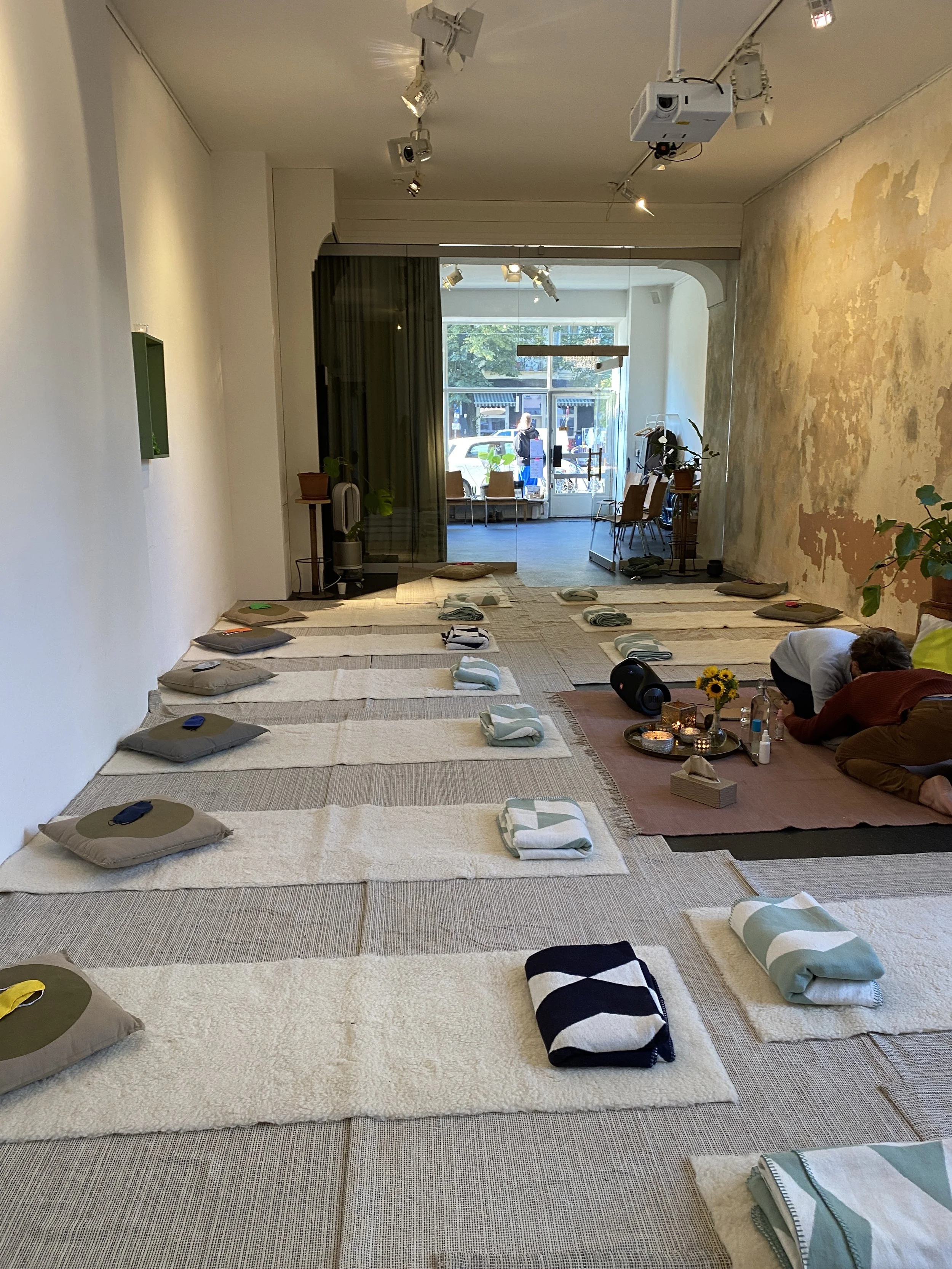 Innenraum eines Raumes mit Sitzgelegenheiten, Decken, Pflanzen und dekorativen Elementen, vorbereitet für eine Sitzung oder Veranstaltung, mit Masken, Decken, Kerzen und einer Person in der Ecke.