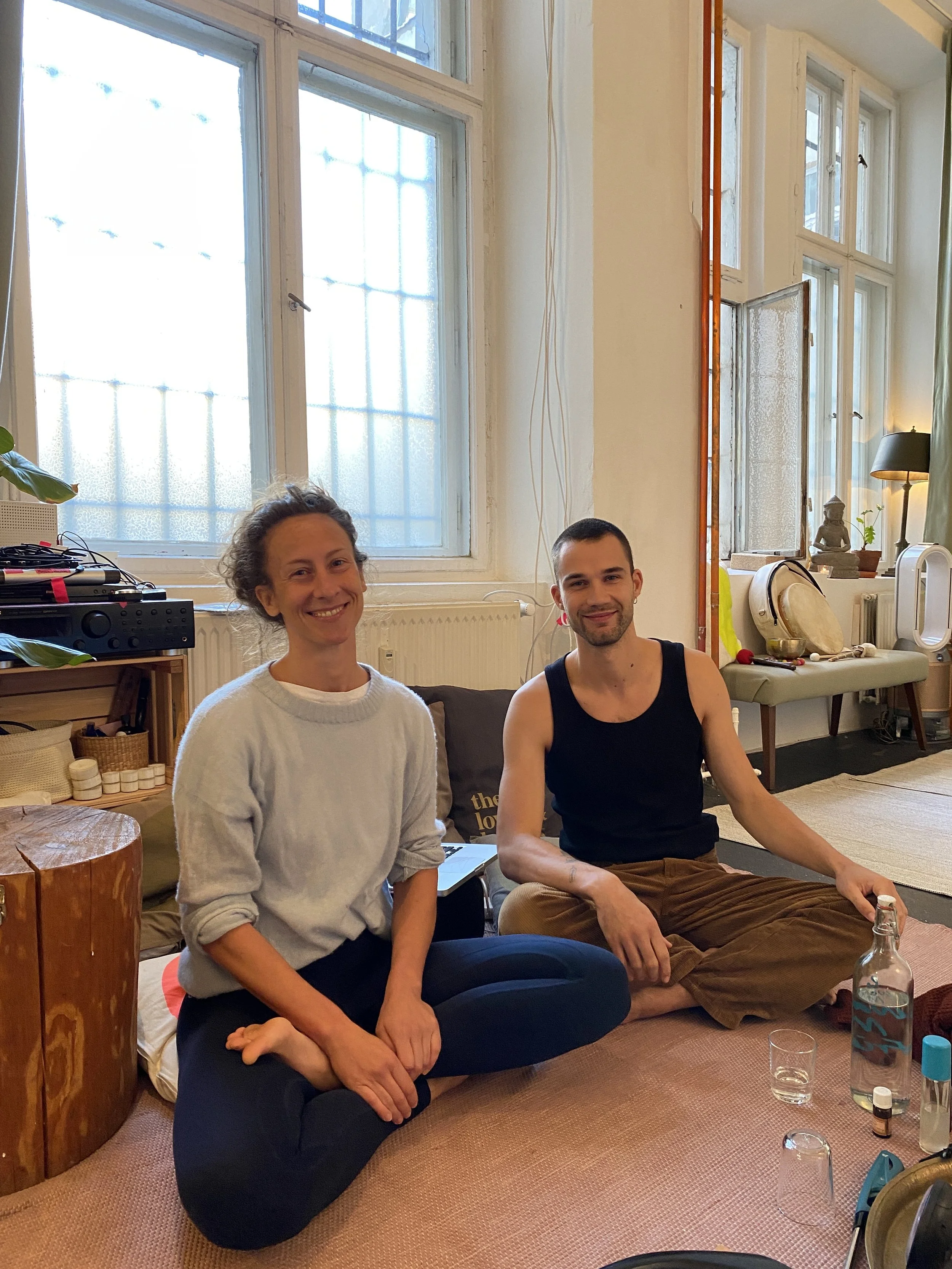 Ein Mann und eine Frau sitzen im Innenraum auf dem Boden, lachen und schauen in die Kamera. Im Hintergrund große Fenster, Dekorationsobjekte und Pflanzen.