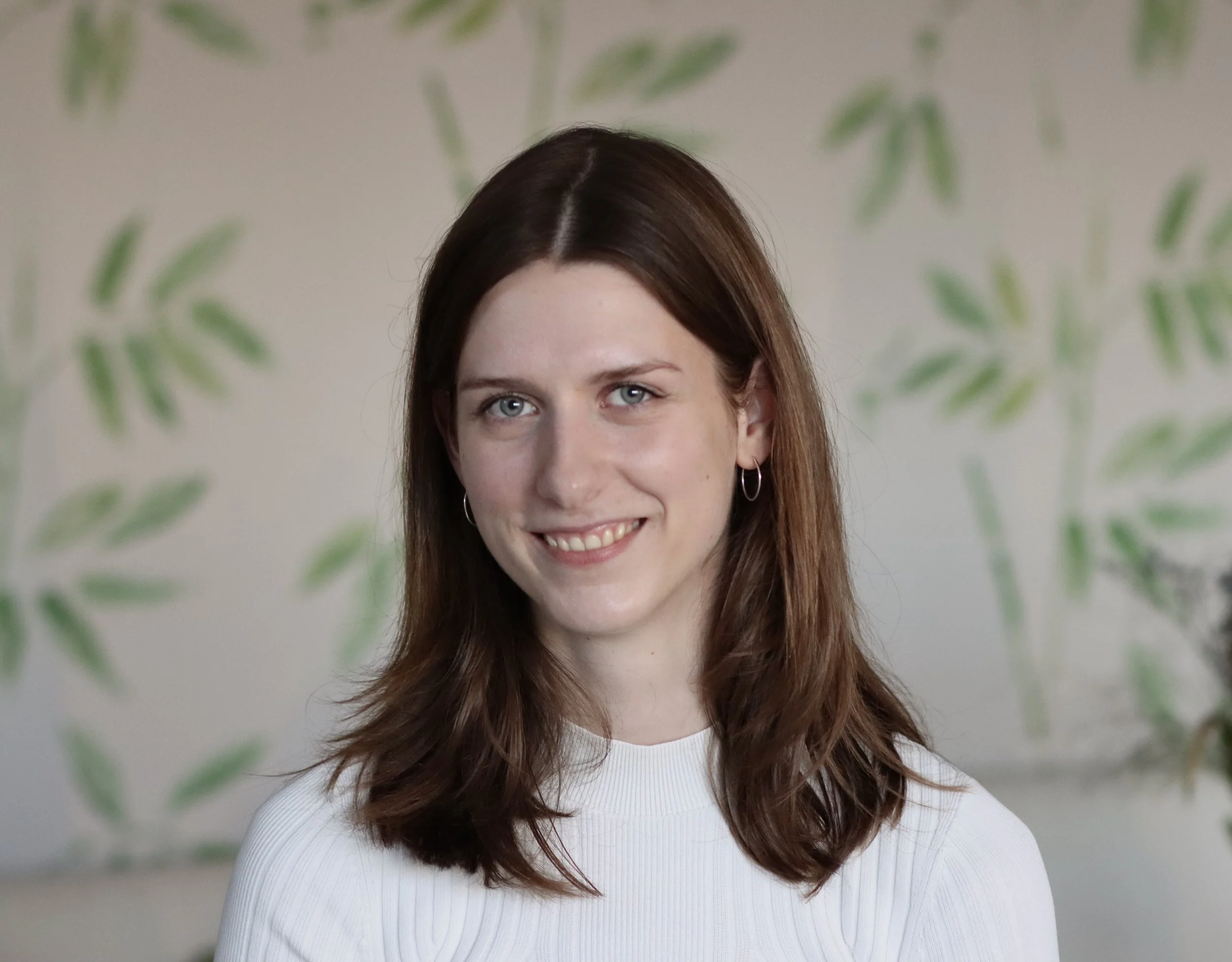 Junge Frau mit braunen, schulterlangen Haaren, trägt weiße Bluse, lächelt vor einer düsteren Wand mit grünen Laubmustern.