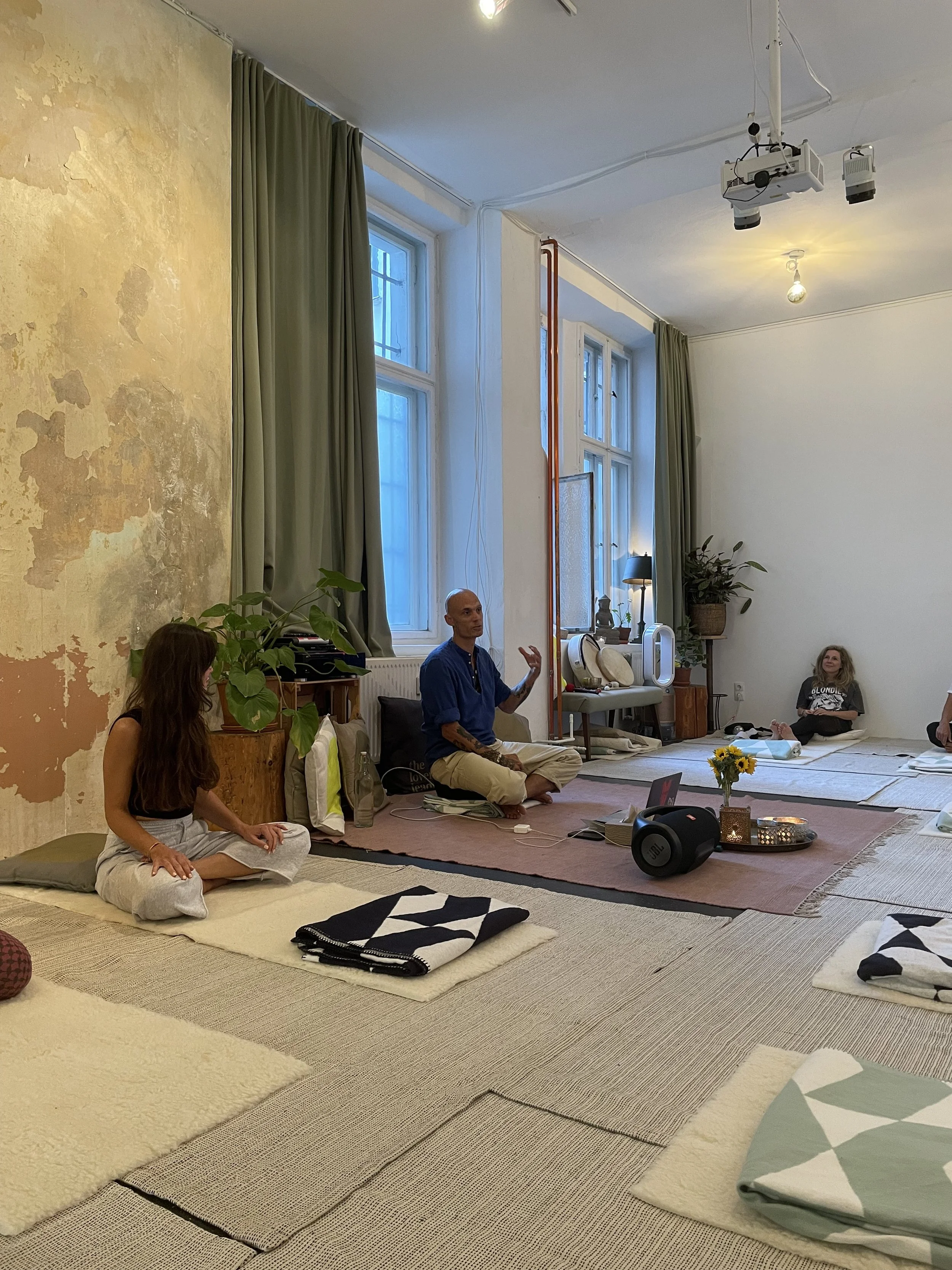 Menschen sitzen in einem gemütlichen Raum auf Teppichen und Kissen, während jemand spricht und schwingt.