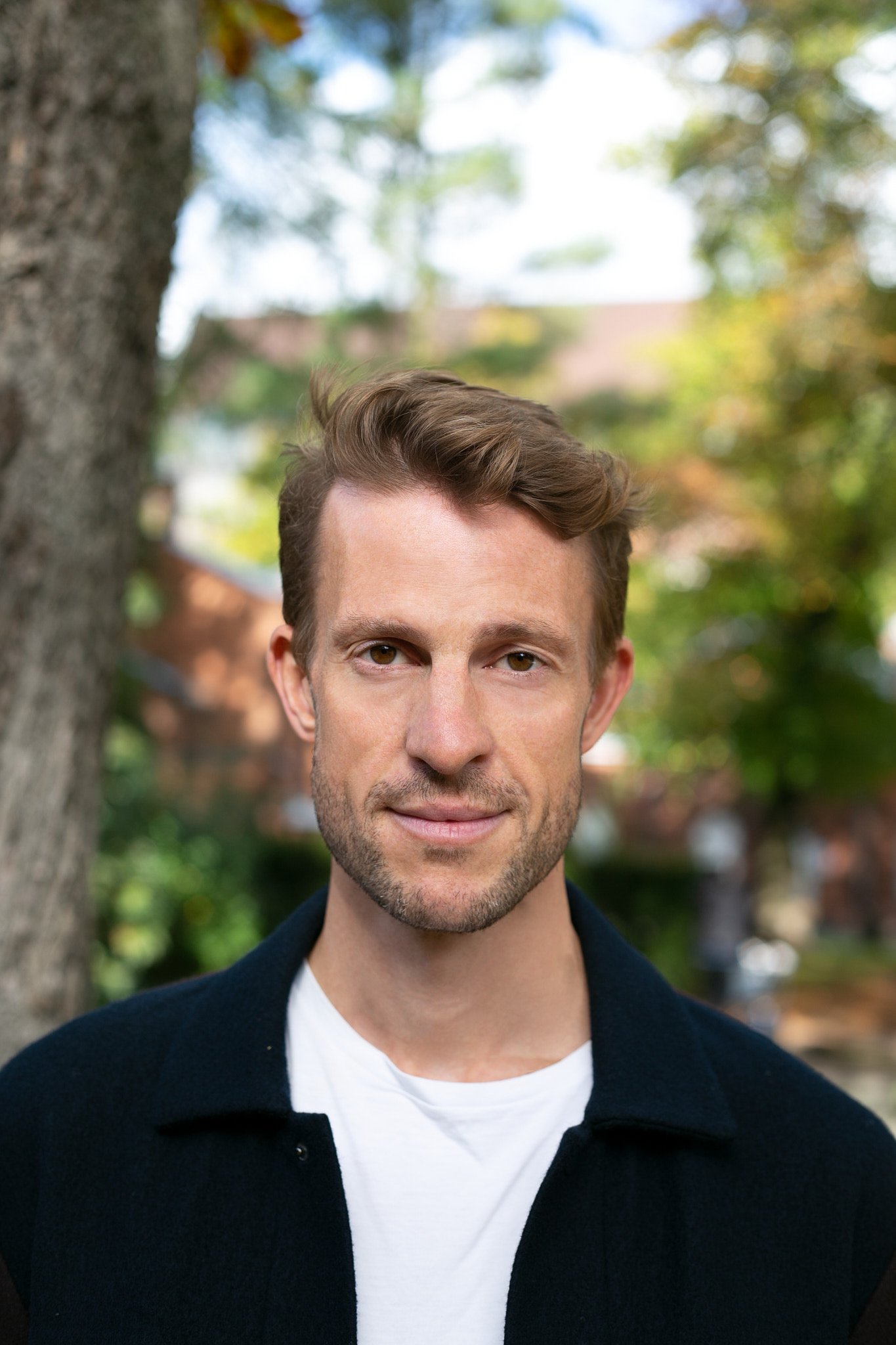 Ein Mann mit braunen Haaren und Bart posiert vor einem Baum im Freien, im Hintergrund sind Häuser und Bäume sichtbar, bei sonnigem Wetter.