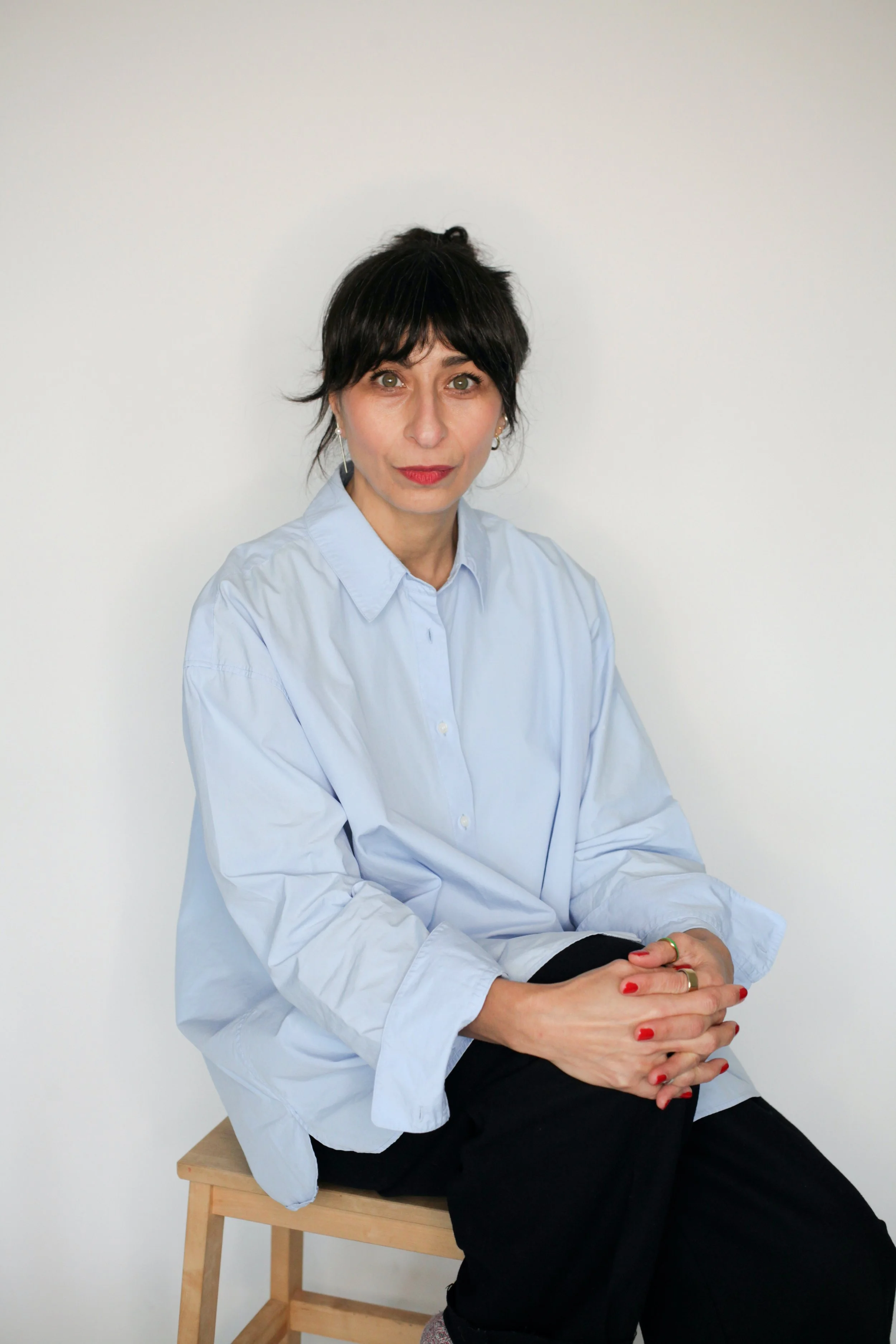 Frau mit schwarzen, kurzen, gewellten Haaren, in einem hellblauen Hemd sitzend auf einem Holzstuhl vor weißem Hintergrund.