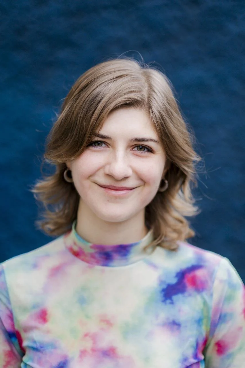 Ein junges Mädchen mit schulterlangem braunem Haar, lächelnd vor einem blauen Hintergrund, trägt ein farbenfrohes, paisleyartiges Turtleneck.