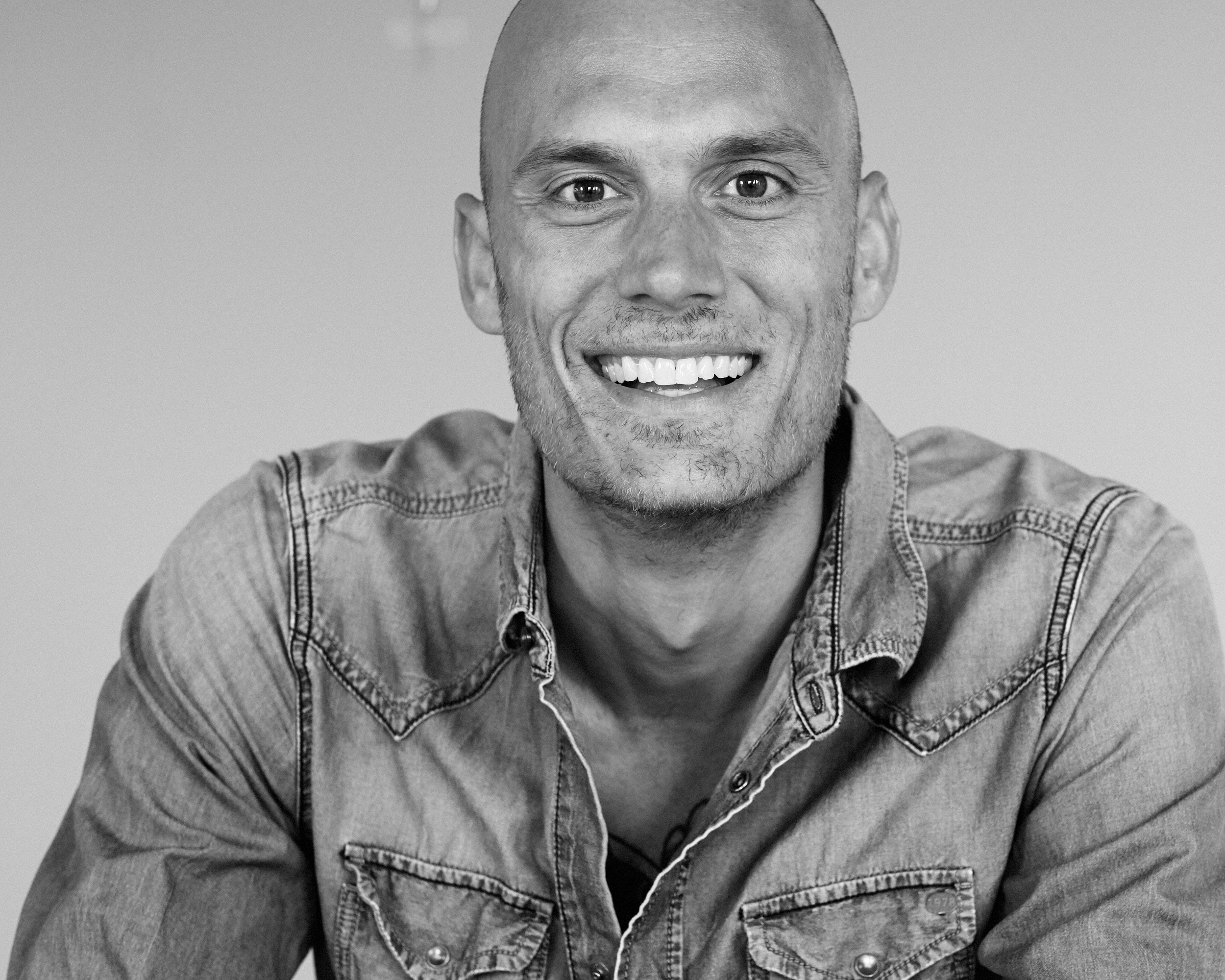 Schwarz-weiß-Fotografie eines lachenden Mannes mit Locken und einem gestreiften Hemd.