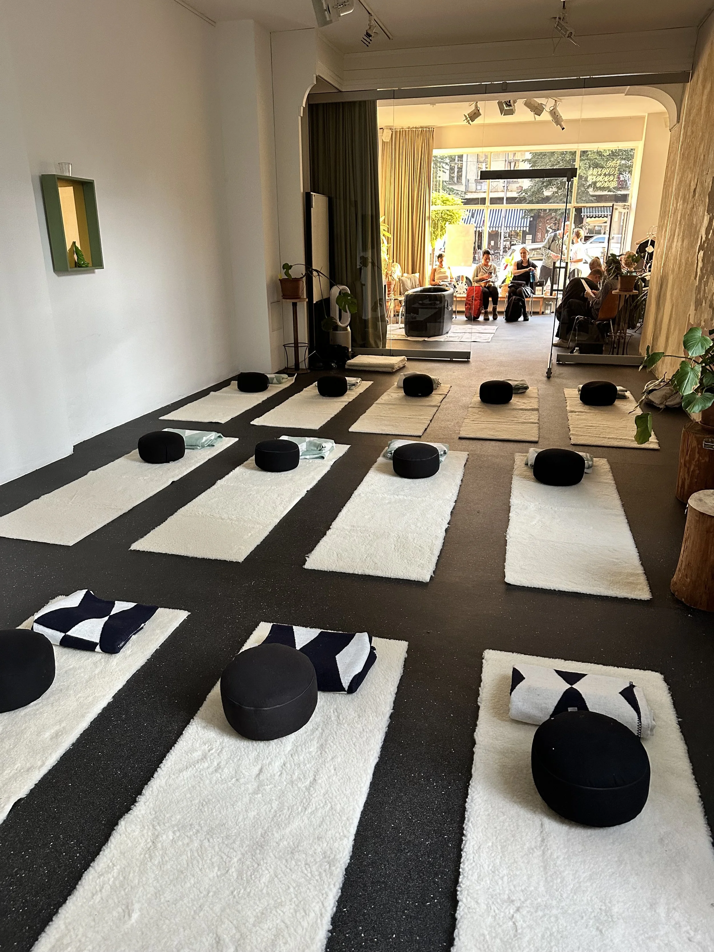 Innenaufnahme eines Yoga-Studios mit auf den weißen Matten und schwarzen Kissen angeordneten Yogakissen und Handtüchern. Im Hintergrund sieht man die Fenster, durch die Sonnenlicht in den Raum fällt, und eine Gruppe von Menschen, die sich vor dem Ein