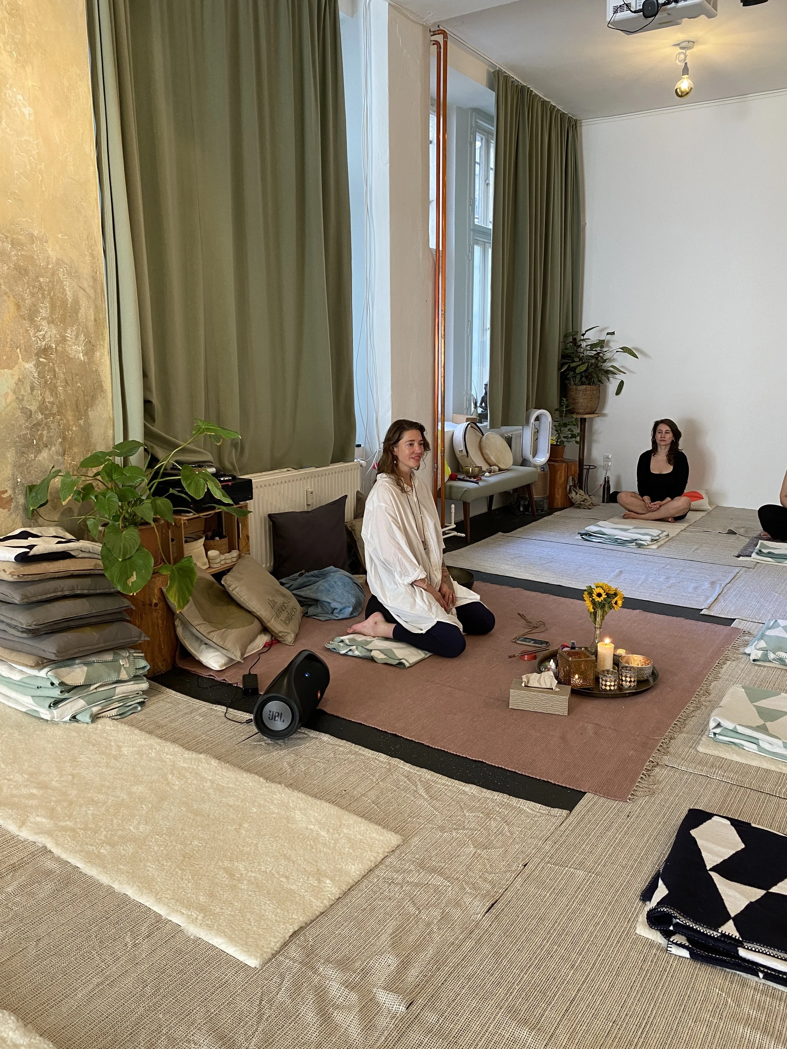 Zwei Frauen sitzen auf Teppichen in einem Raum mit hohen Fenstern, grünen Vorhängen und Pflanzen, umgeben von Decken, Kissen, einer Kerze, einer Blume und einigen Gegenständen für eine spirituelle Praxis.
