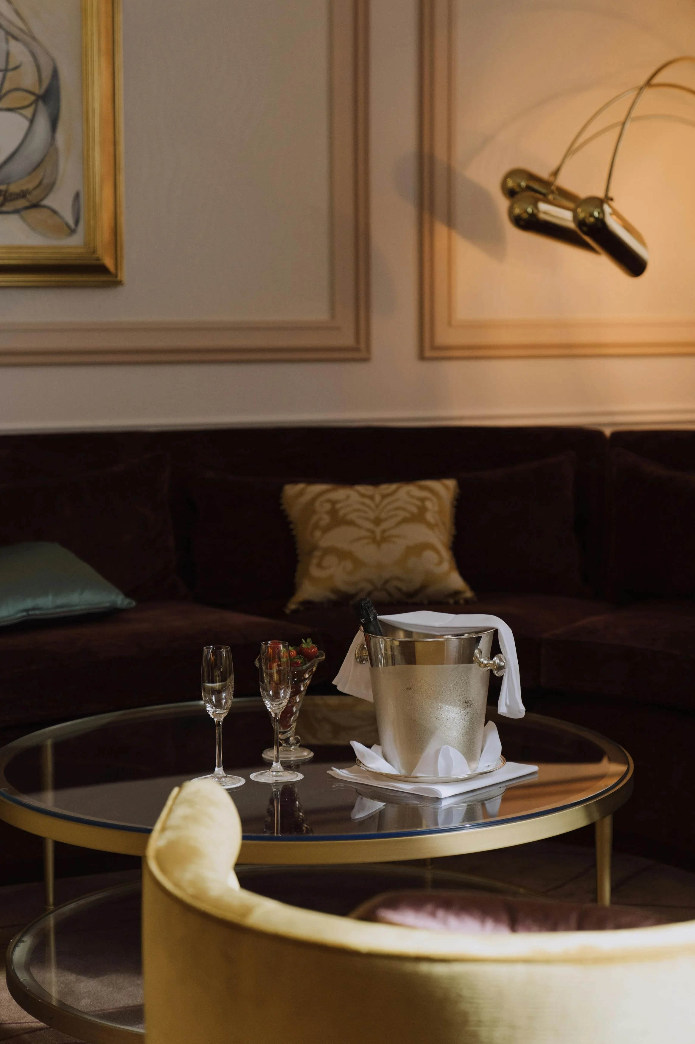 A luxurious living room with a glass coffee table, a champagne bucket with white cloth, two champagne flutes, a bowl of strawberries, a velvet sofa with decorative pillows, and a gold-framed wall art and lamp.