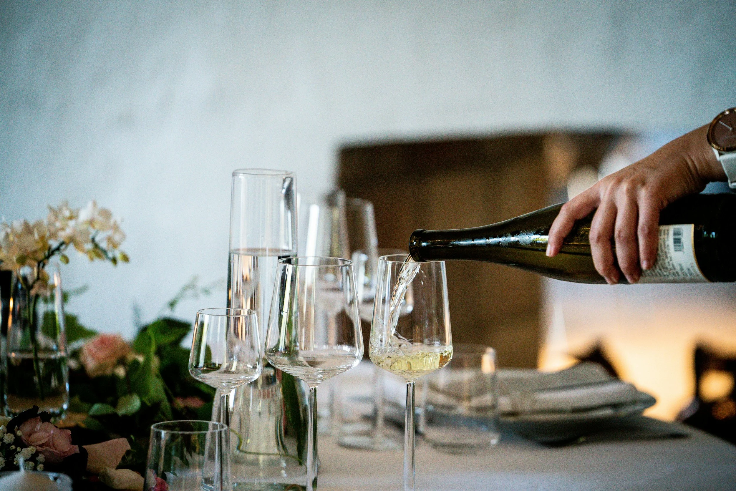 Someone pouring white wine into a glass at a table set with various wine glasses, plates, utensils, and floral arrangements.