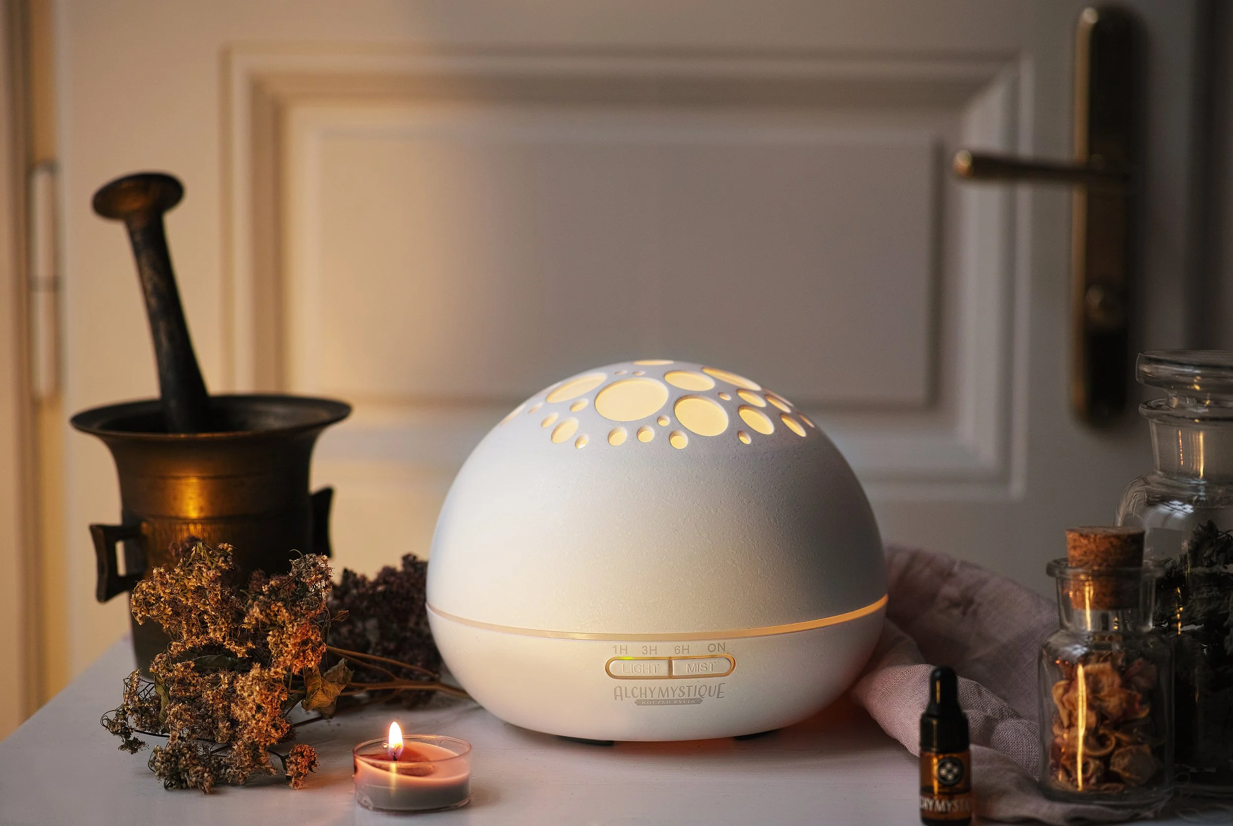 Bílý aromadifuzér Alchymystique se světelným vzorem, obklopený bylinkami a svíčkou v jemném interiérovém světle, produktové lifestyle fotografie.